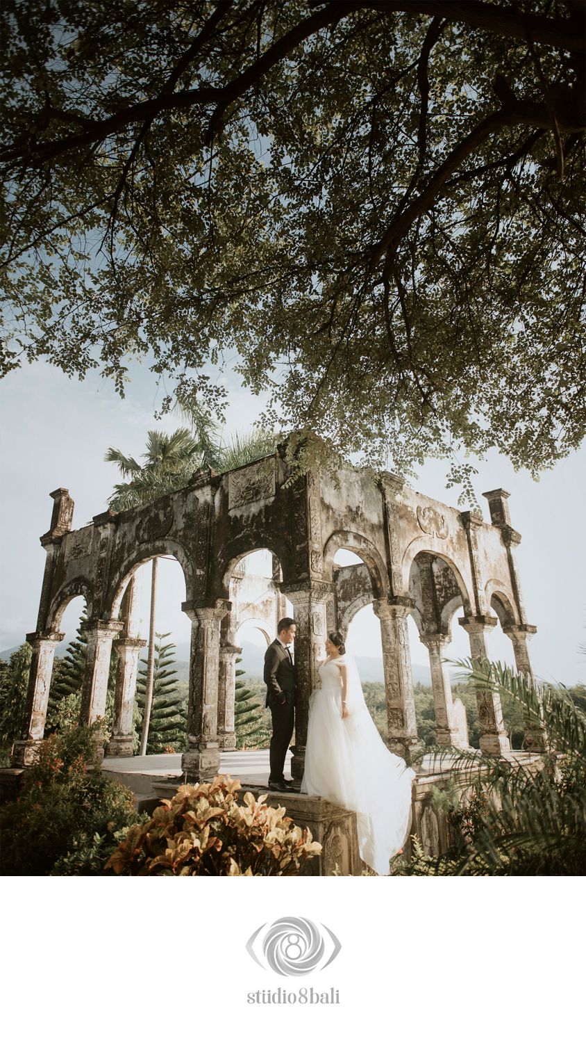 Couple poses romantically at ancient stone arch ruins surrounded by lush tropical trees in a stunning Bali wedding photo.