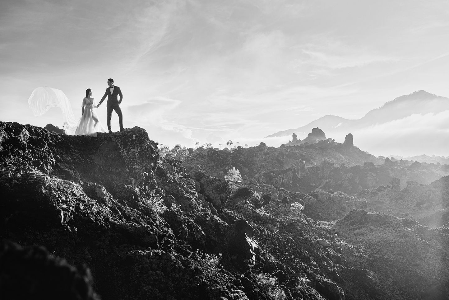 Black and white photo of a couple standing on a rocky volcanic landscape with misty mountains in the background.
