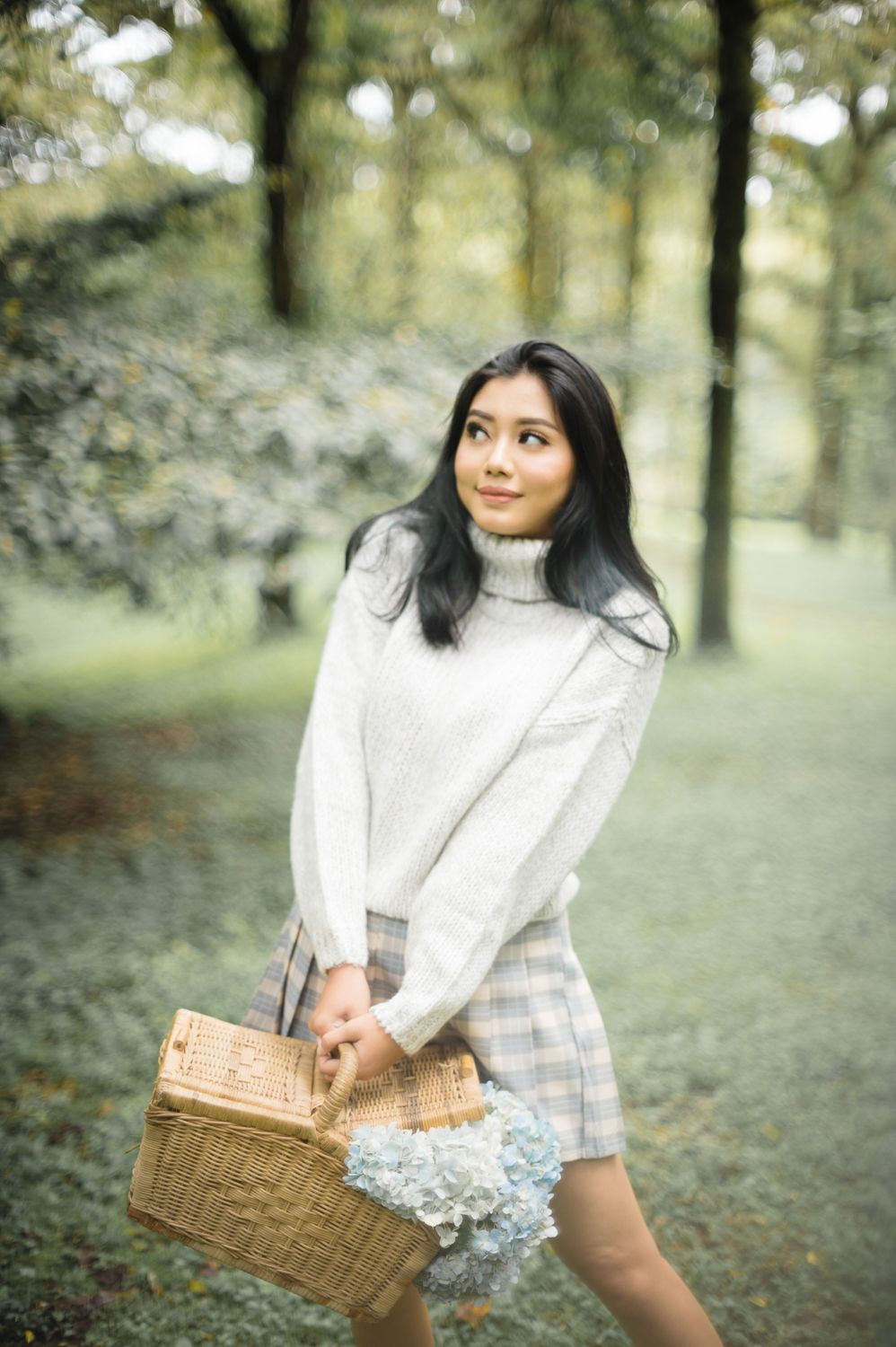 Young woman in cream turtleneck sweater holding wicker basket with flowers in a lush green park setting.