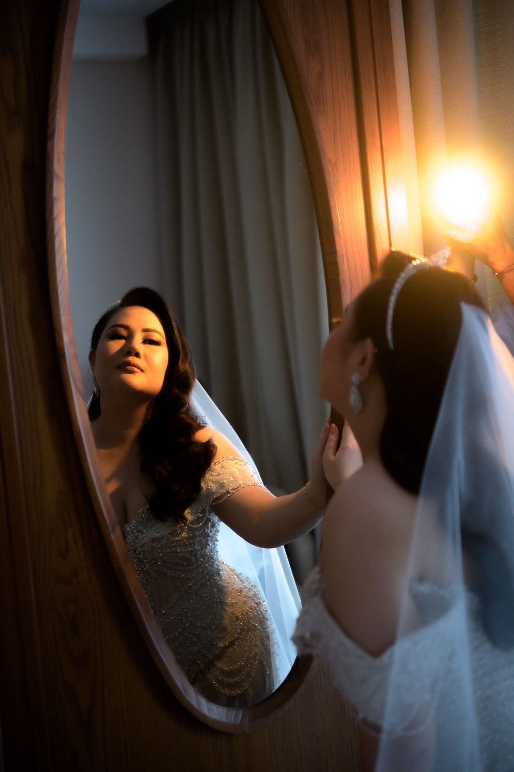 Bride in lace gown adjusts veil while gazing at her reflection in an oval mirror, warmly lit by a vintage bulb.