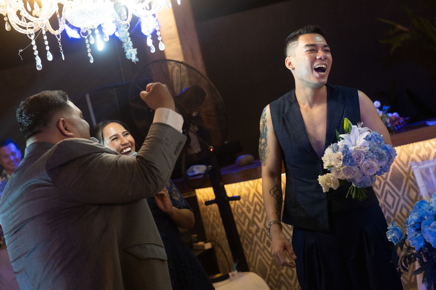 Joyful man with tattoos laughing and holding blue flowers at a wedding celebration with guests in elegant venue.