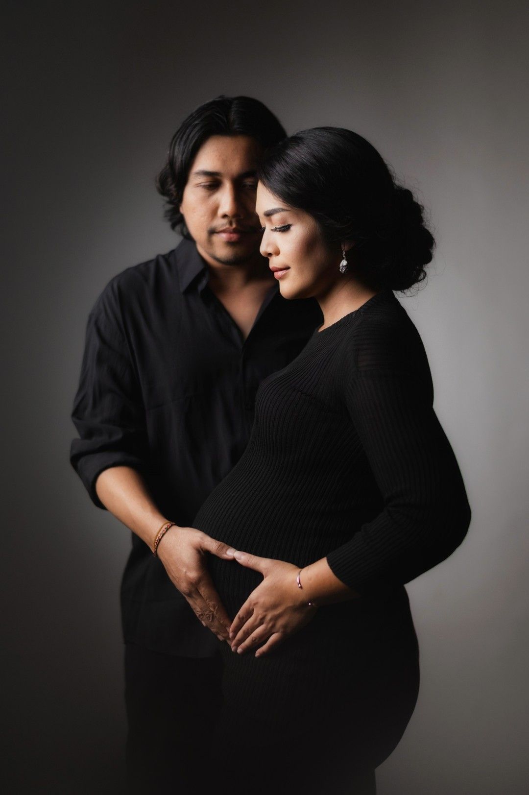 A couple in black outfits sharing a tender maternity portrait moment, hands gently cradling a baby bump.