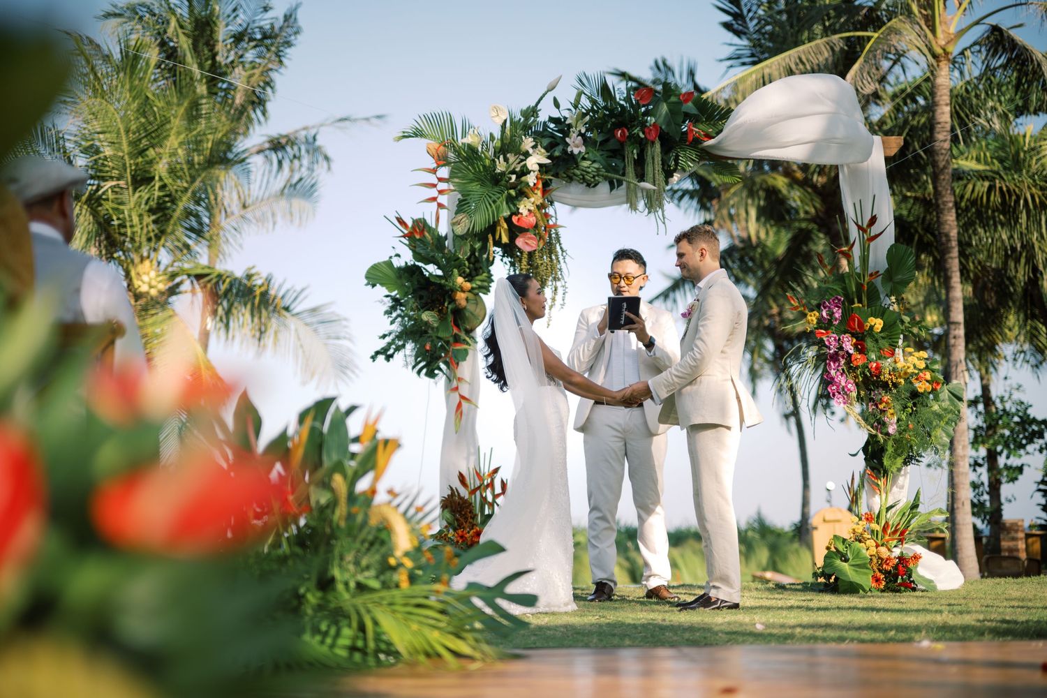 Two grooms exchange vows under a tropical floral arch at an outdoor beachside wedding ceremony.