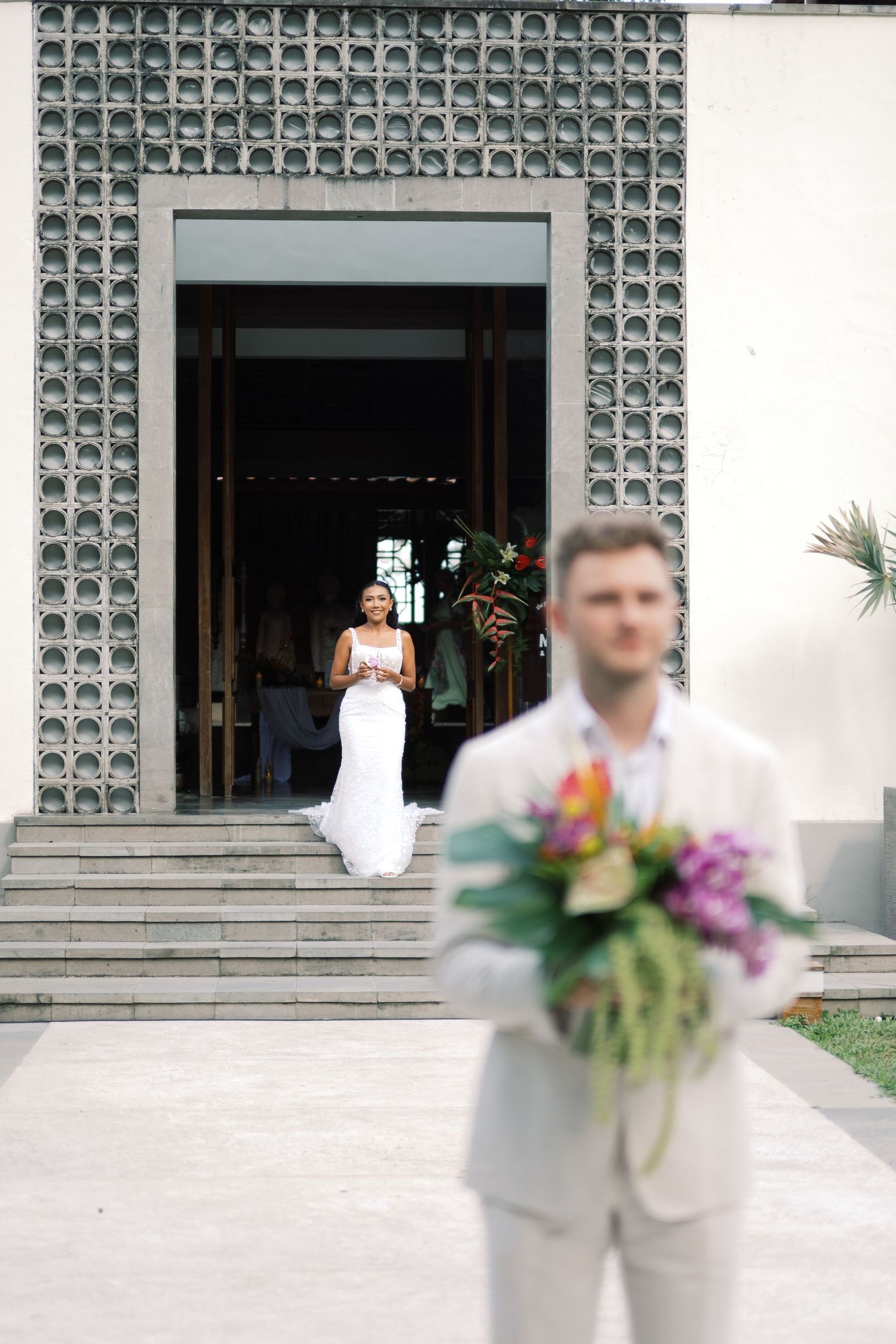 Bride in fitted white gown descends steps toward groom holding tropical bouquet at venue entrance.
