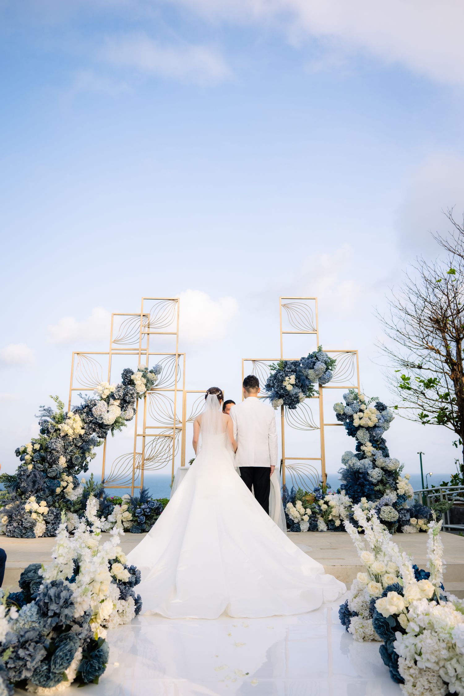 Bride in a long white gown and groom in a white jacket stand facing gold geometric arches with blue and white flowers at an ocean wedding.
