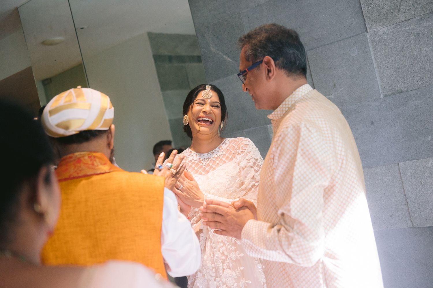 A woman in white lace laughs joyfully with guests in traditional Indian attire at a festive celebration.