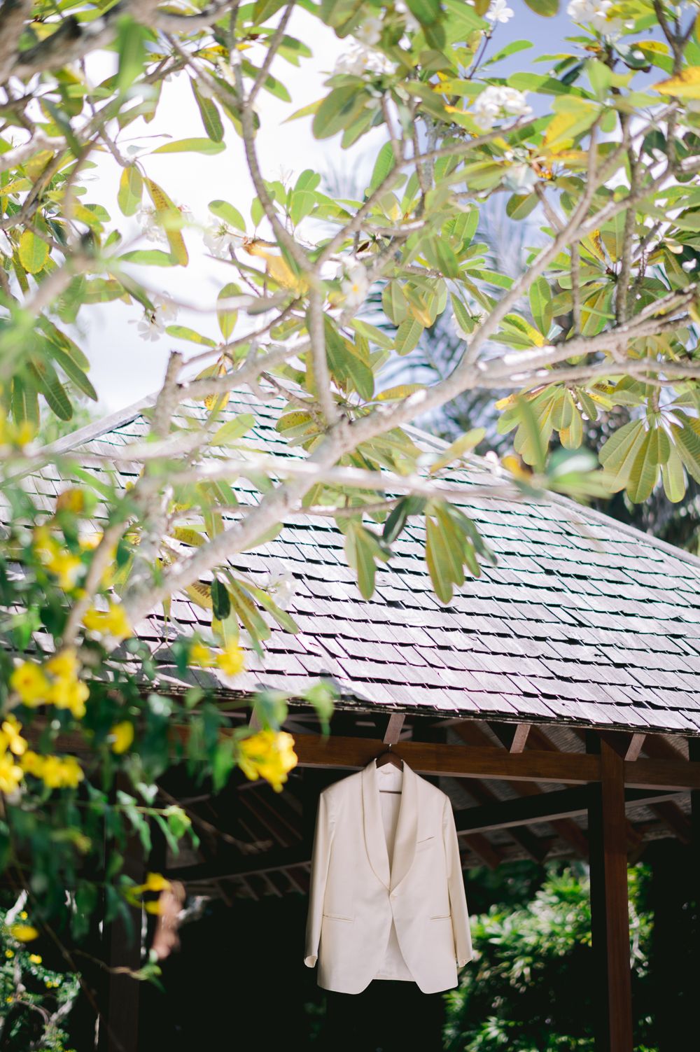 White blazer hanging on a wooden gazebo surrounded by tropical trees with yellow flowers.