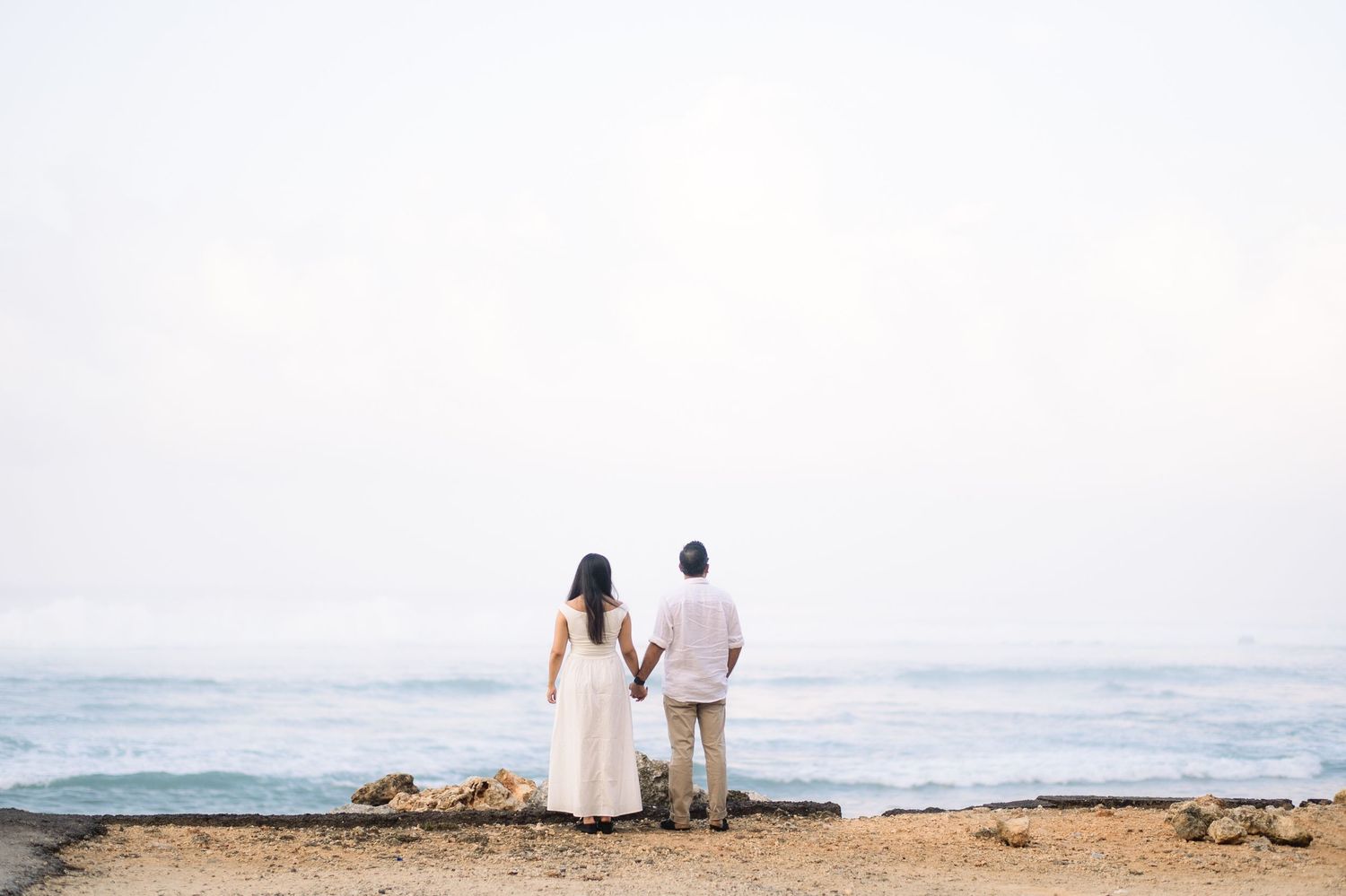 Couple holding hands facing the ocean on a rocky beach shore in matching white and light pink outfits.