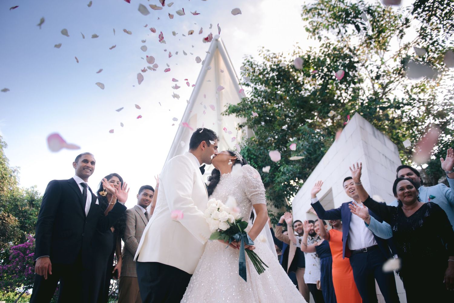 Newlywed couple kissing outdoors as guests toss flower petals in celebration at a church wedding ceremony.