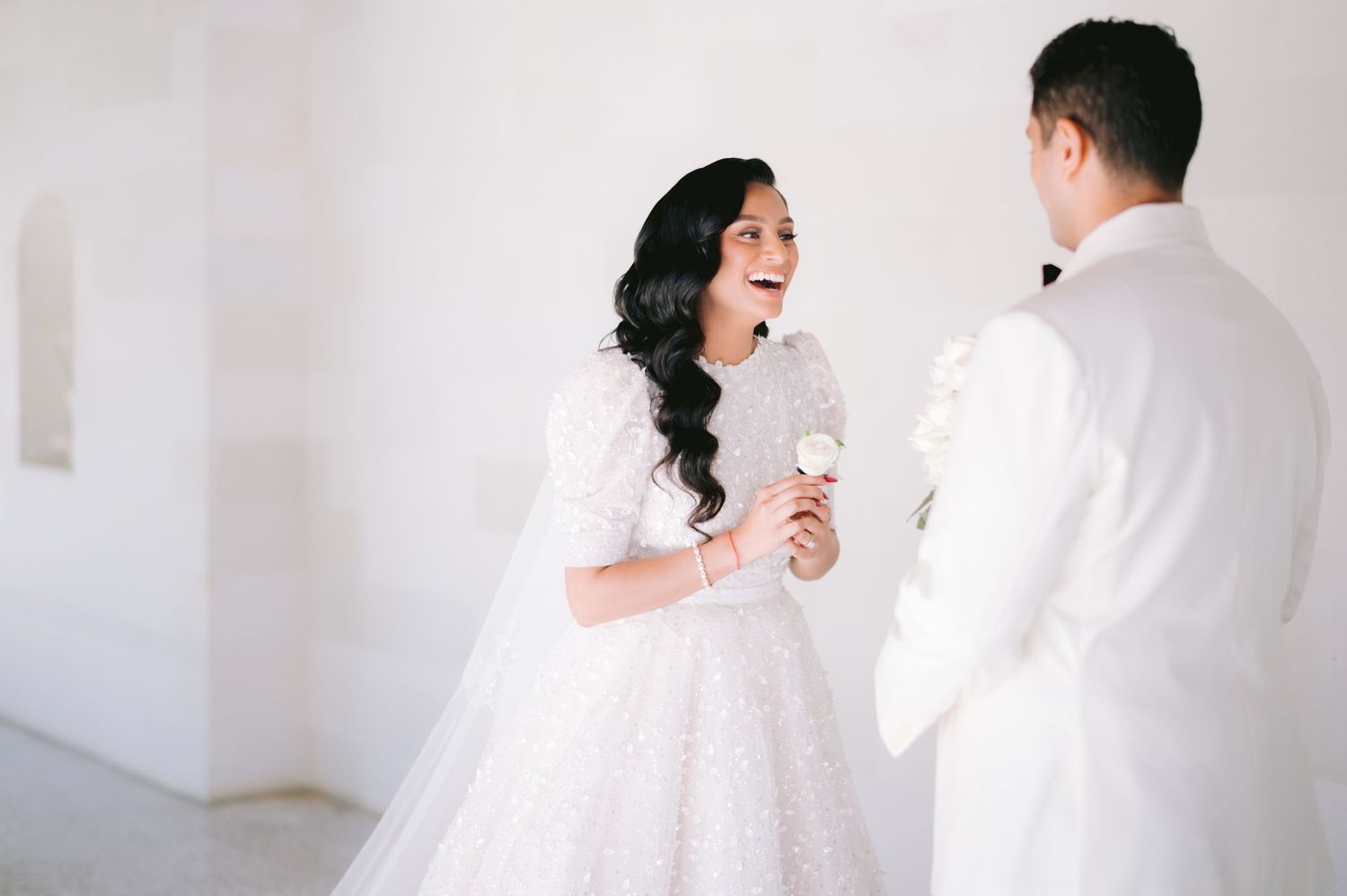 Bride in white lace gown laughing with groom in white tuxedo during elegant wedding ceremony.