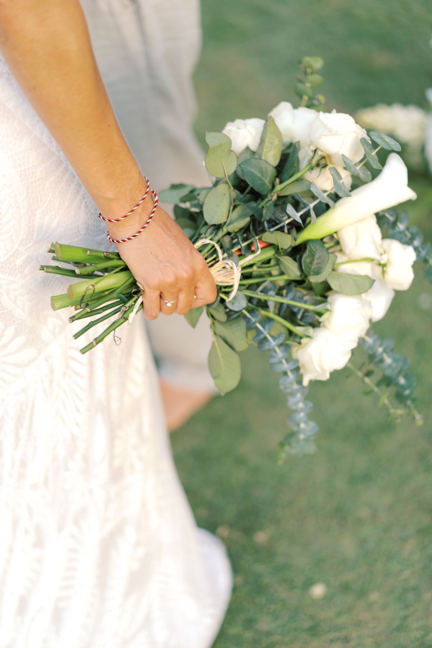 Bride holding a white floral bouquet with greenery and calla lilies, wearing a lace dress and delicate bracelets.
