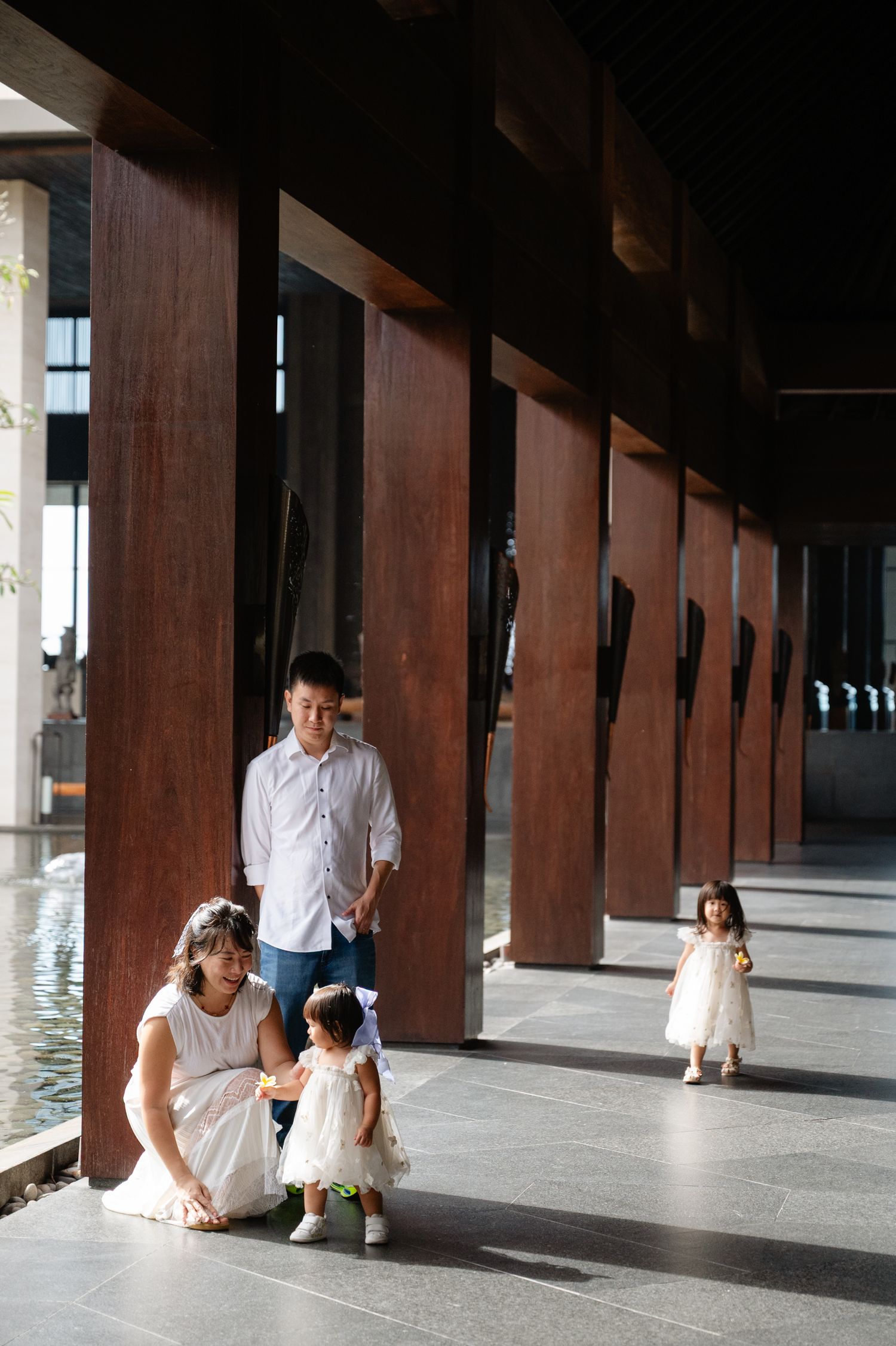 Asian family with two young girls in white dresses posing in a modern building corridor with wooden pillars.