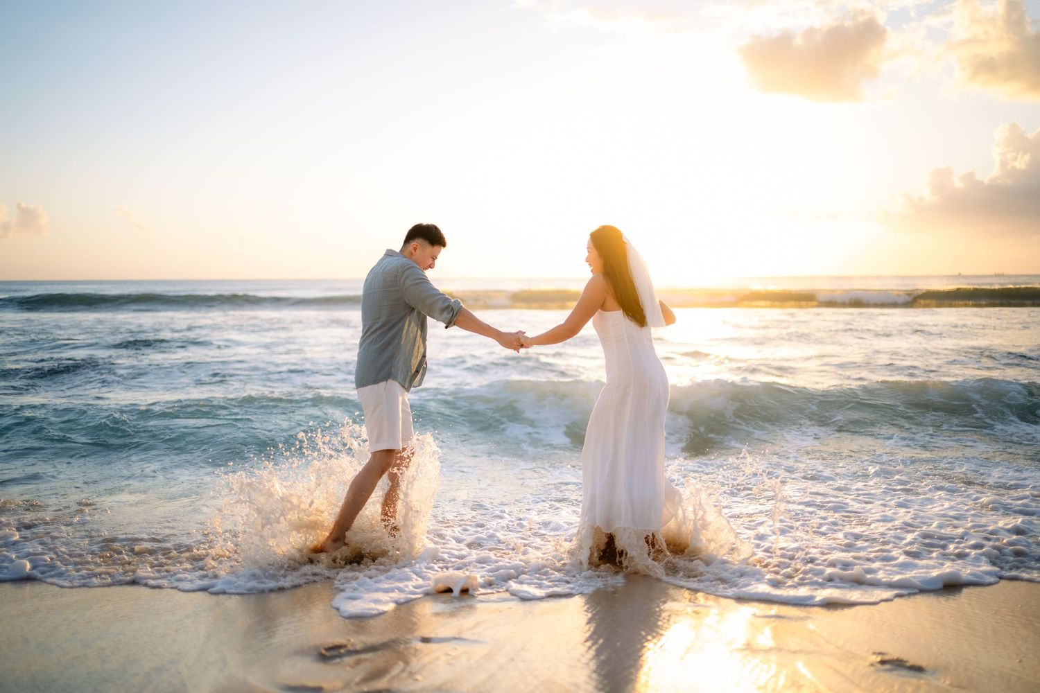 Couple holding hands dancing in ocean waves at sunset on a tropical beach.
