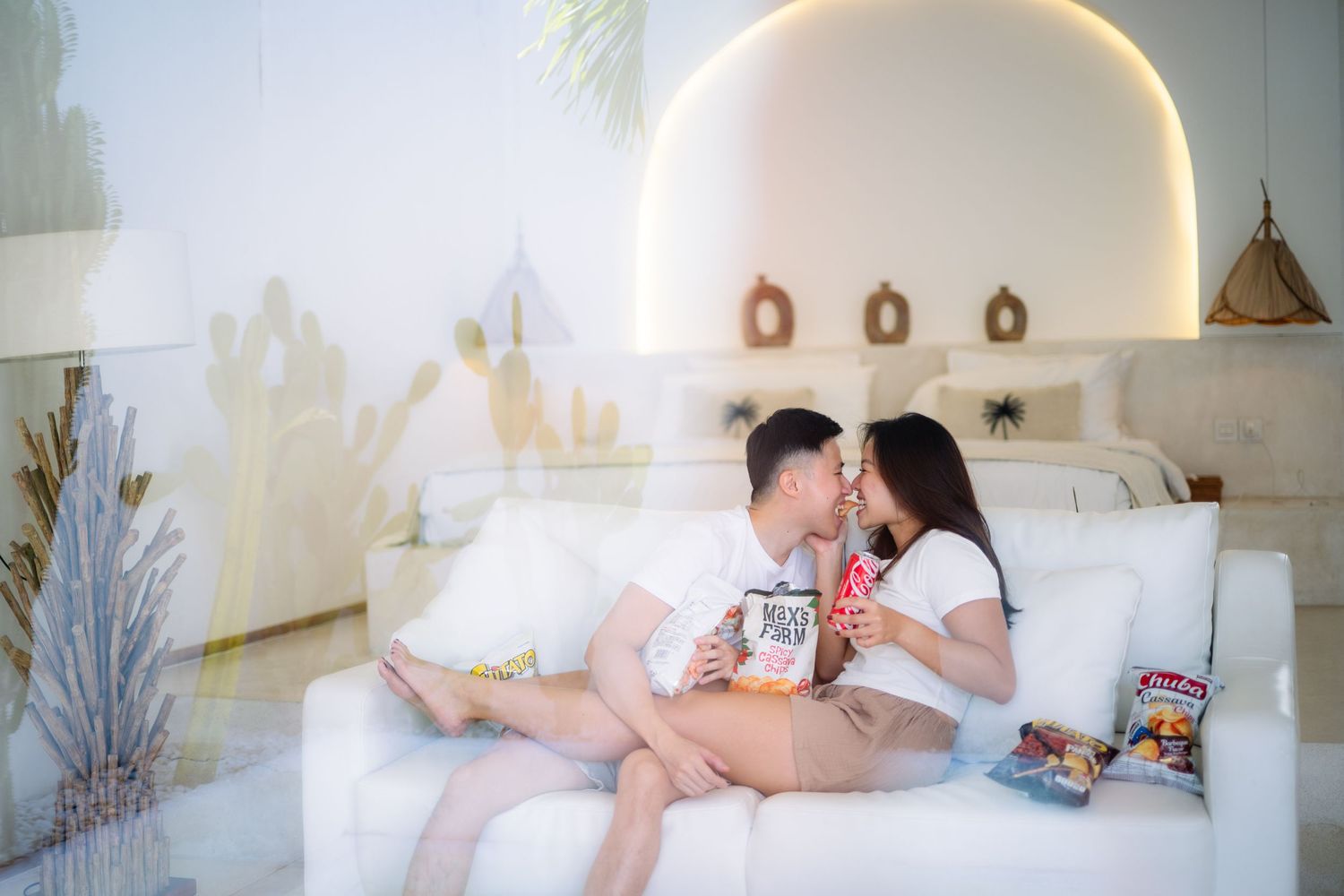 Couple sharing a romantic kiss on a white sofa in a stylish bright bedroom with snacks nearby.