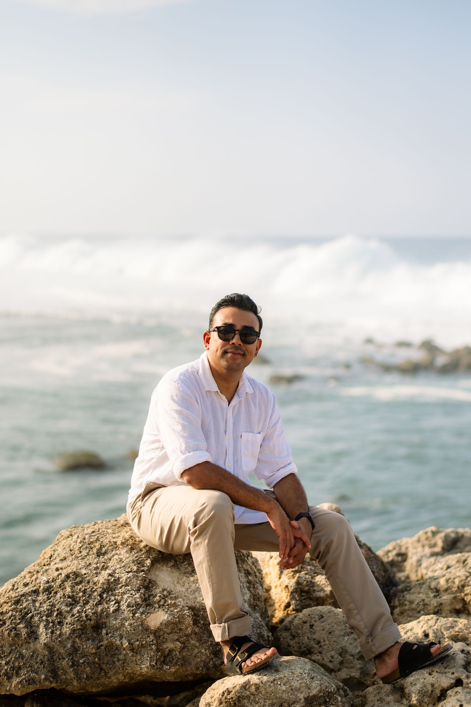 Man in white shirt and sunglasses sitting on coastal rocks with ocean waves in the background.