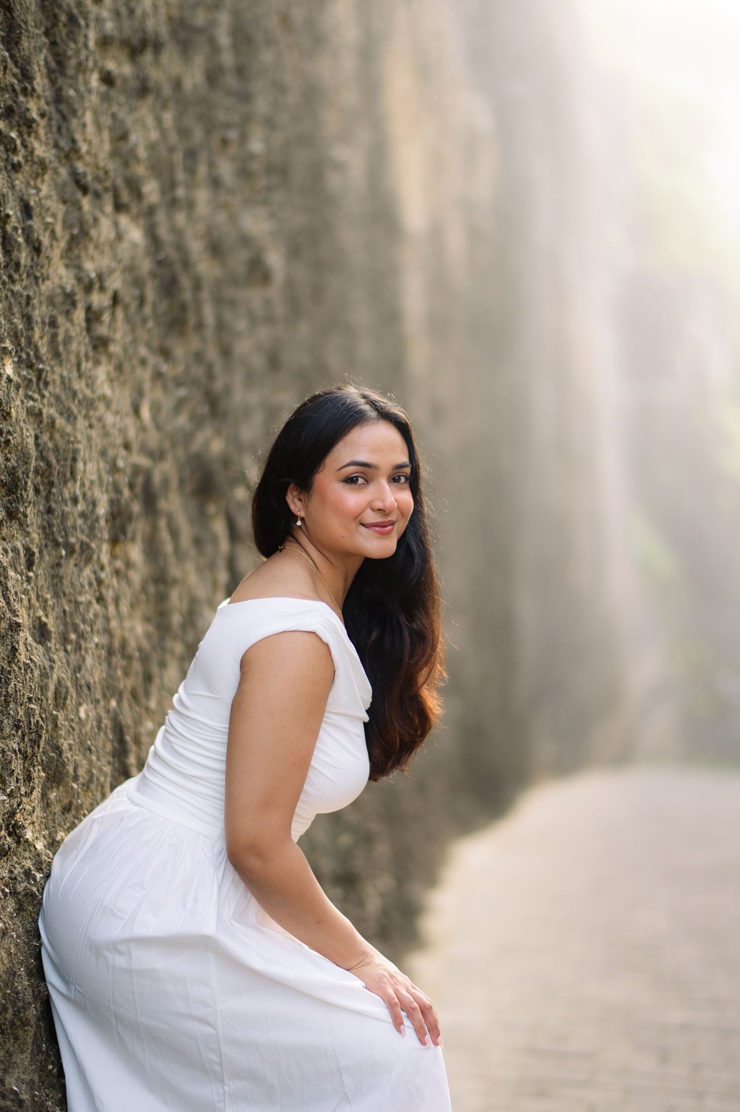 A woman in a white off-shoulder dress leans against a stone wall, smiling gracefully in a sunlit narrow pathway.