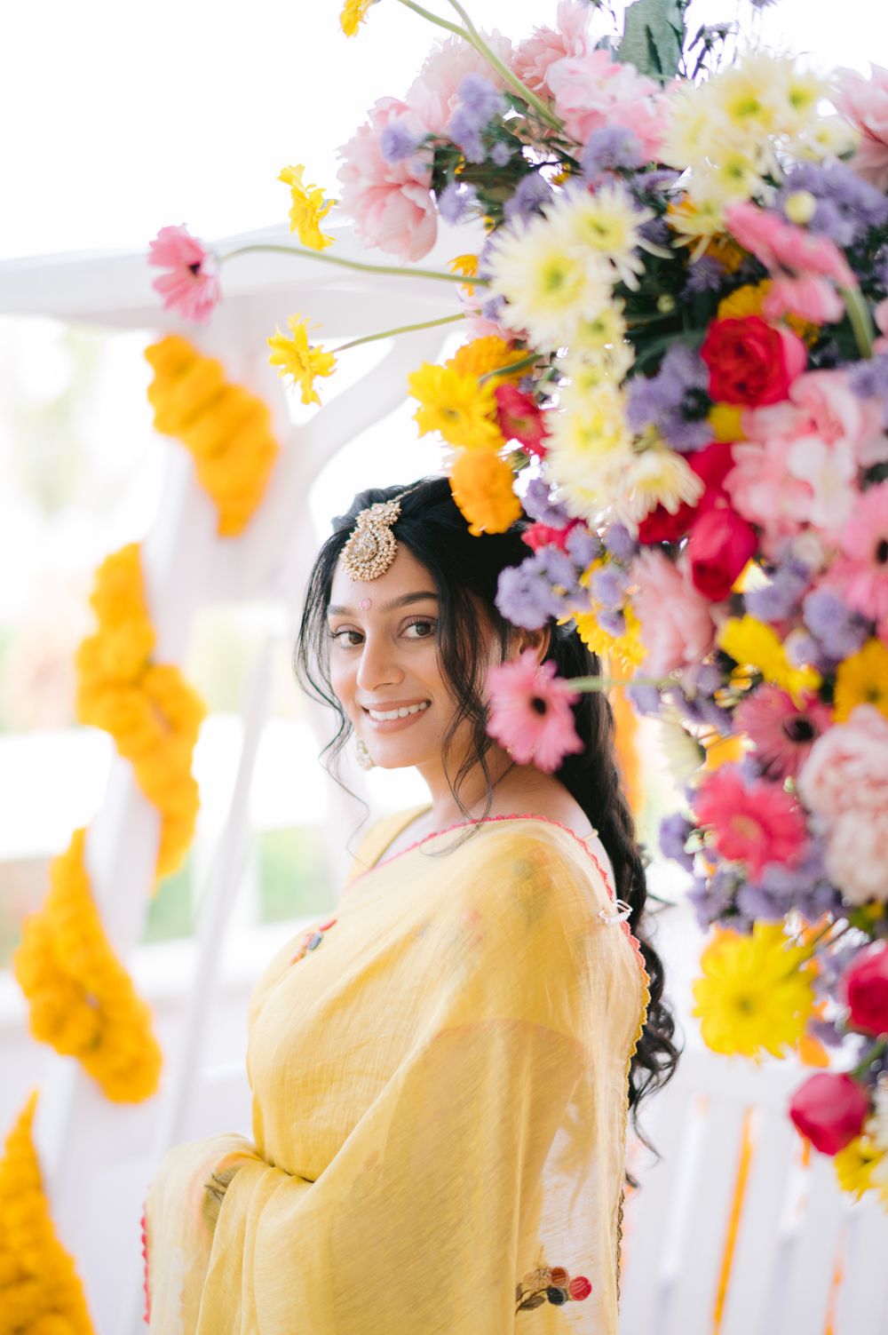 Smiling bride in yellow saree with maang tikka poses before colorful floral arch at Indian wedding celebration.