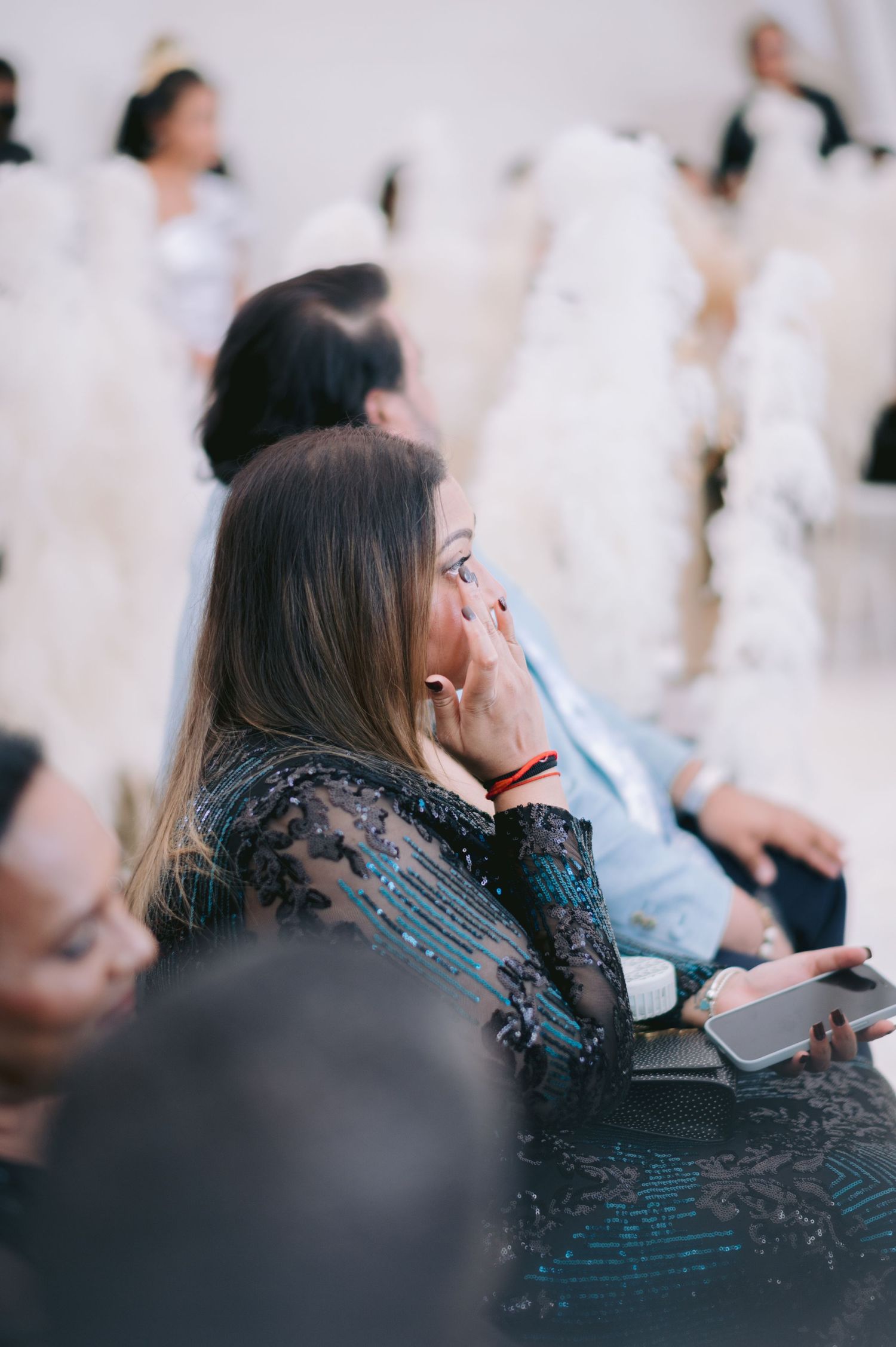 Emotional wedding guest in black lace dress wiping tears, seated among blurred white gowns in elegant venue.