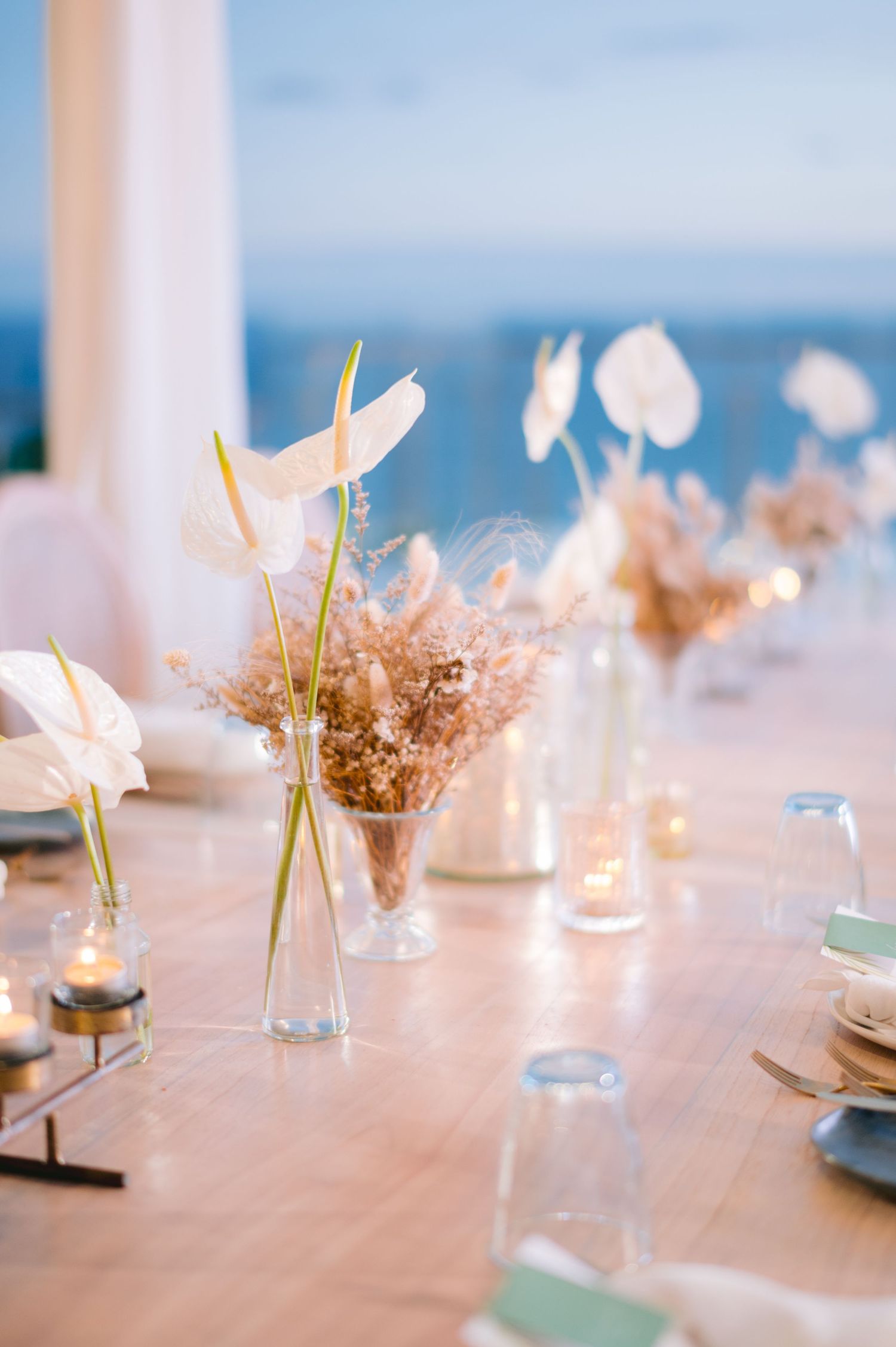 Elegant wedding table with white anthurium flowers, dried botanicals in glass vases, and candles by the sea.