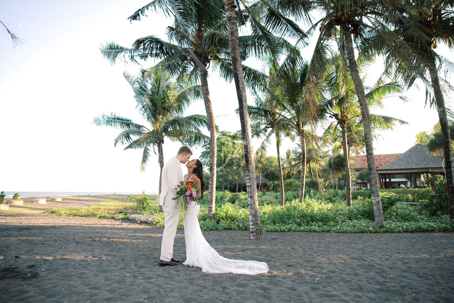 Bride and groom share a romantic kiss on a black sand beach surrounded by tall palm trees at a tropical wedding.