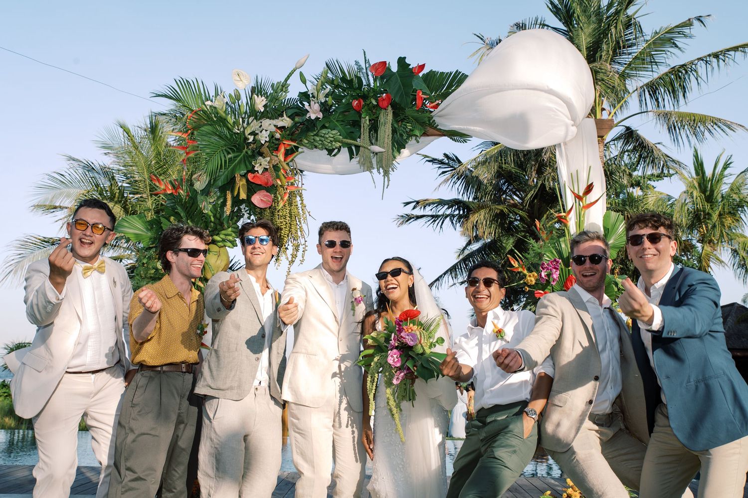 Bride and groom celebrate with groomsmen under floral arch at a tropical beach wedding.