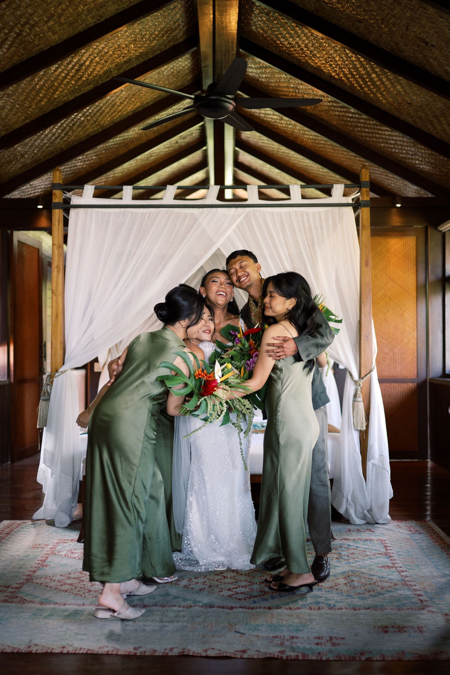 Bride in white gown embraces bridesmaids in green satin dresses inside a Balinese-style canopy bedroom.