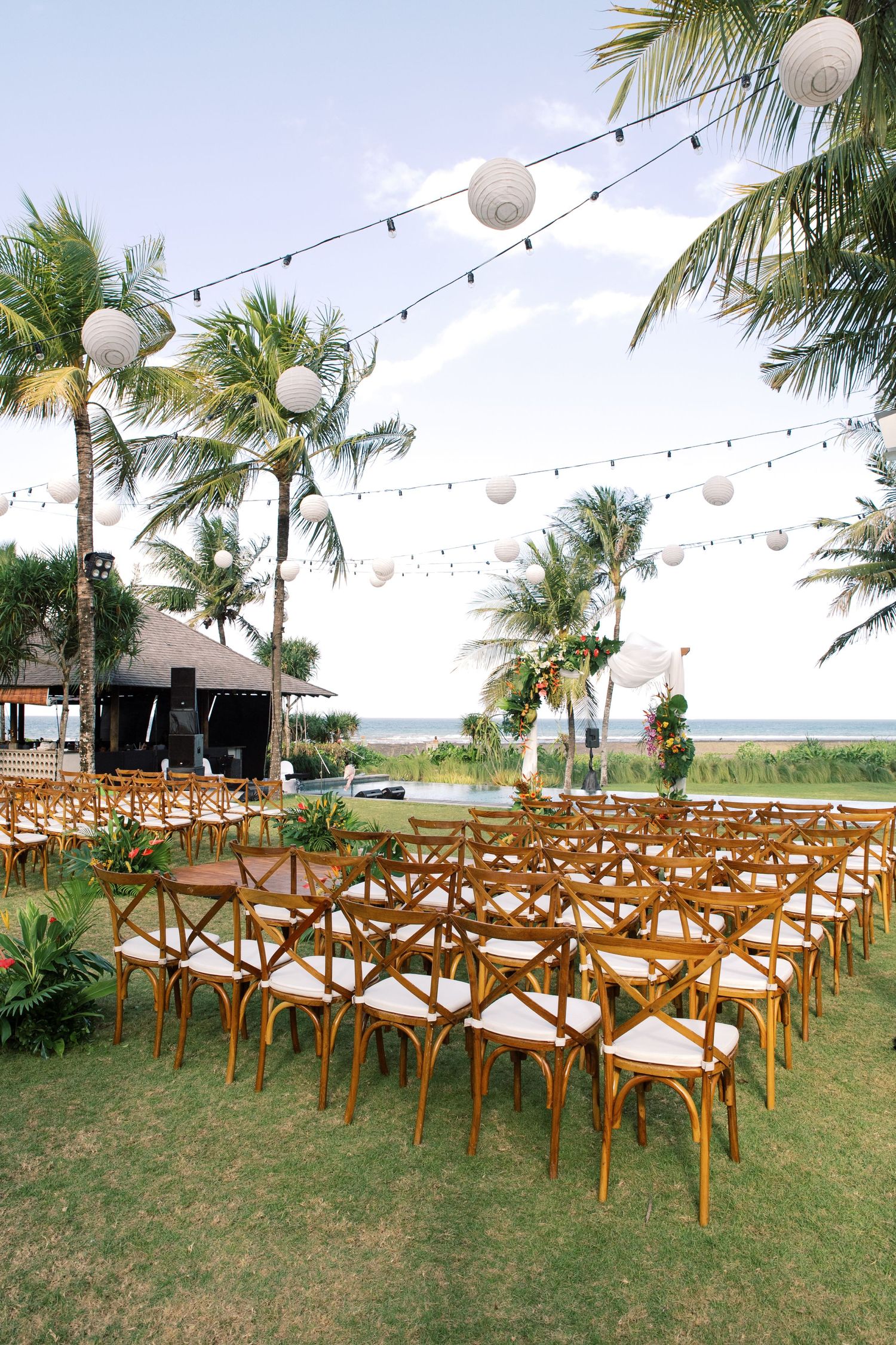 Outdoor beach wedding setup with wooden chairs, palm trees, and white paper lantern decorations.