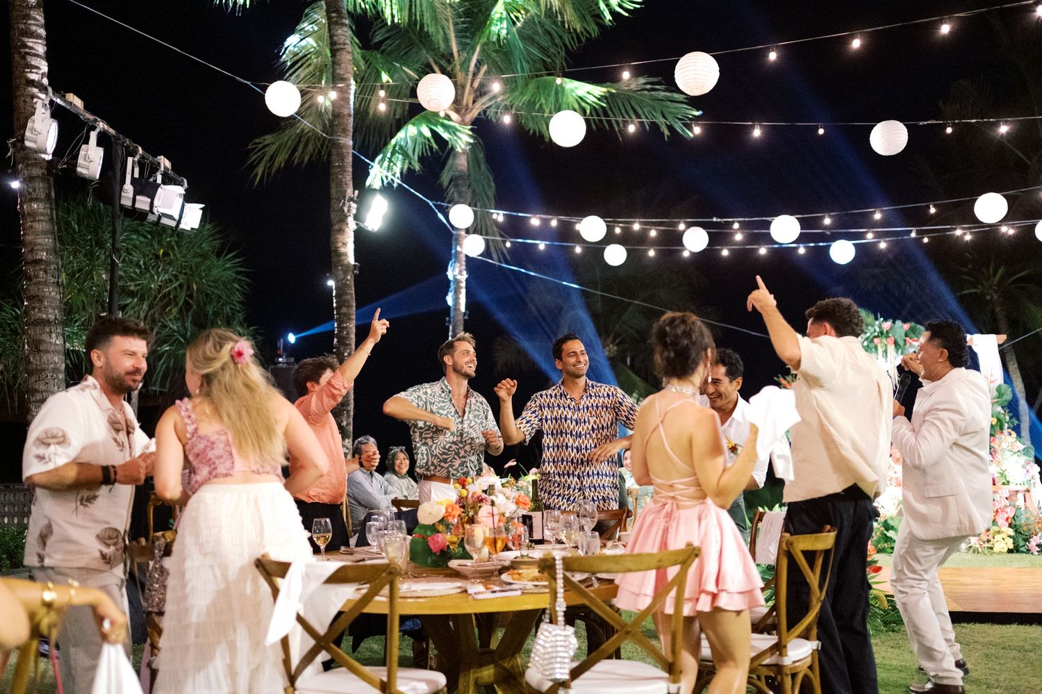 Wedding guests dance and celebrate under string lights at outdoor tropical evening reception.