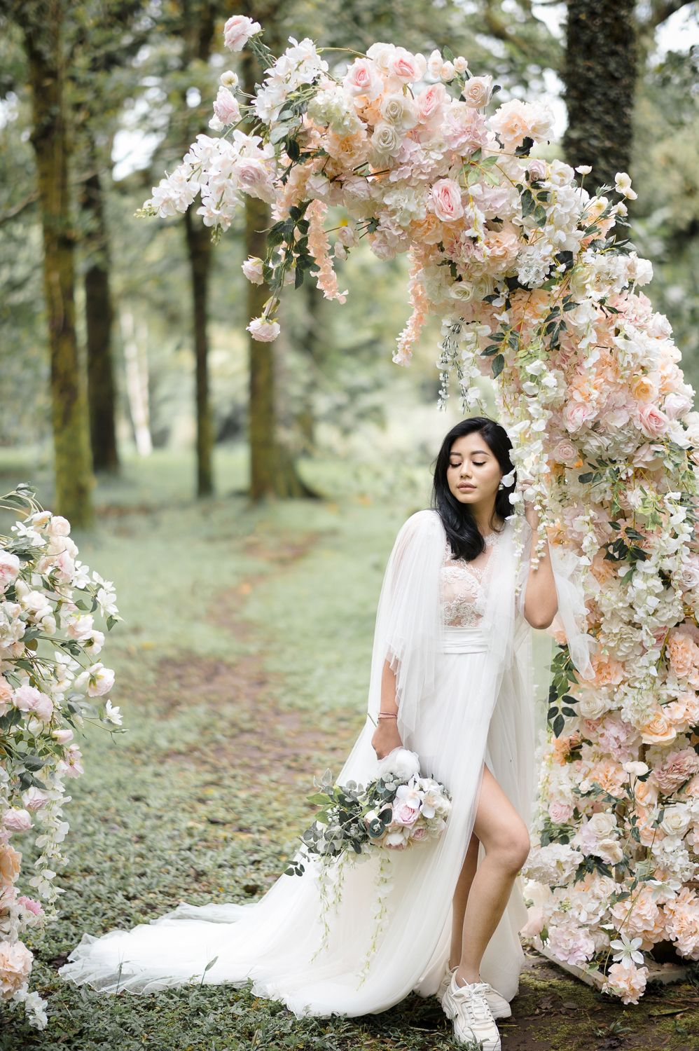 Bride in white gown poses under blush floral arch in forest, holding bouquet in dreamy outdoor wedding.