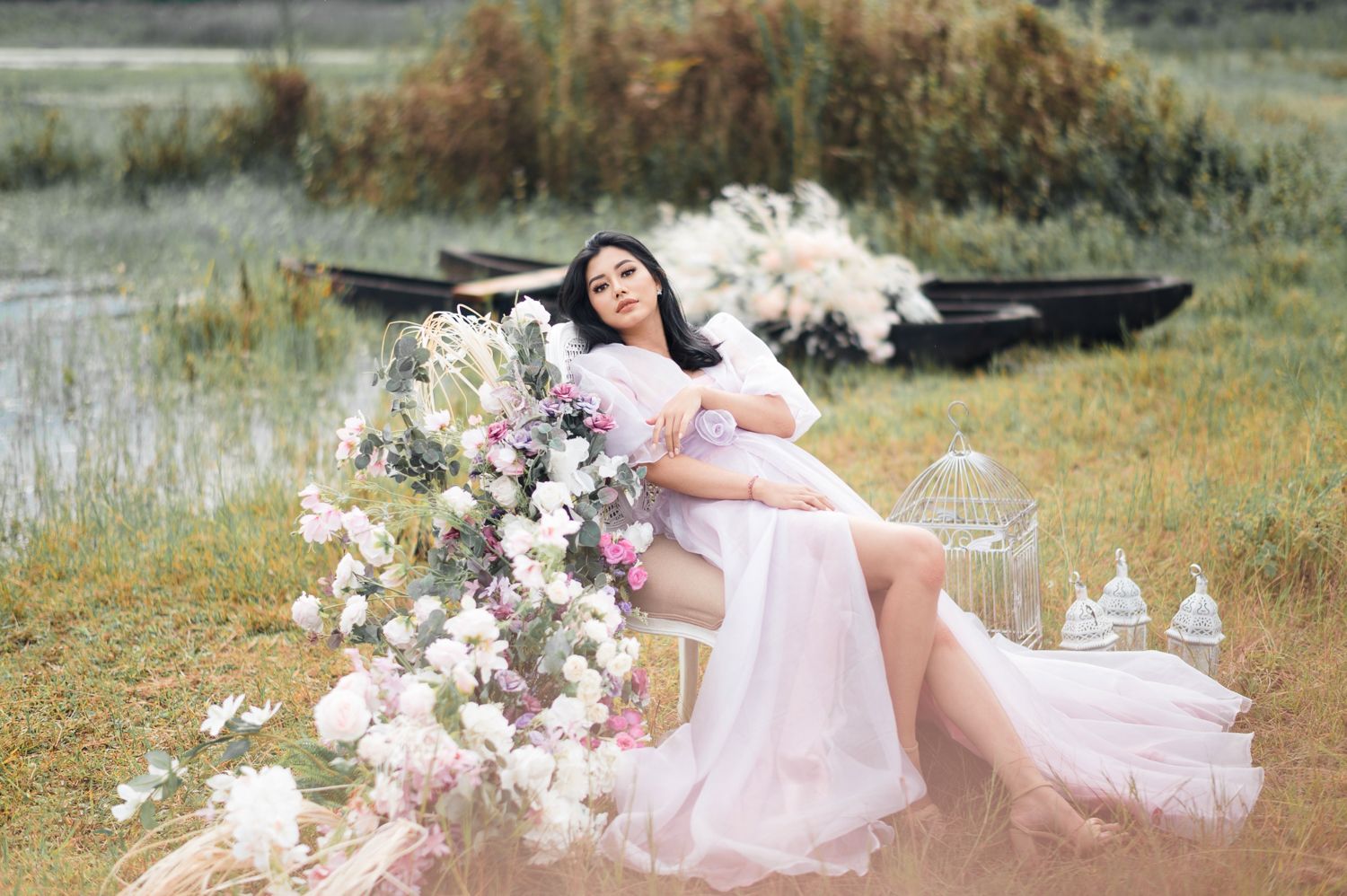 Pregnant woman in pink gown poses with flowers and birdcage by a serene lake with a wooden boat backdrop.
