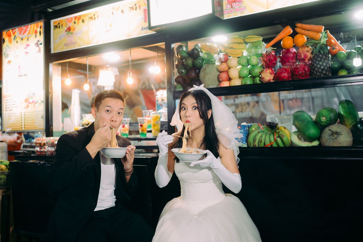 Bride and groom in wedding attire eating noodles at a colorful outdoor food market stall.