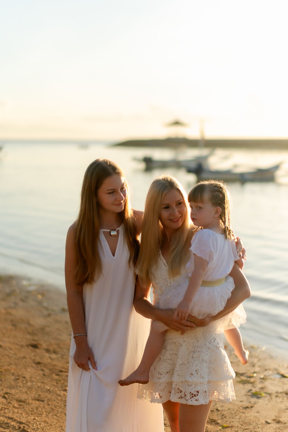 Mother holding young daughter while teenage girl stands beside them on a sunny beach at sunset.