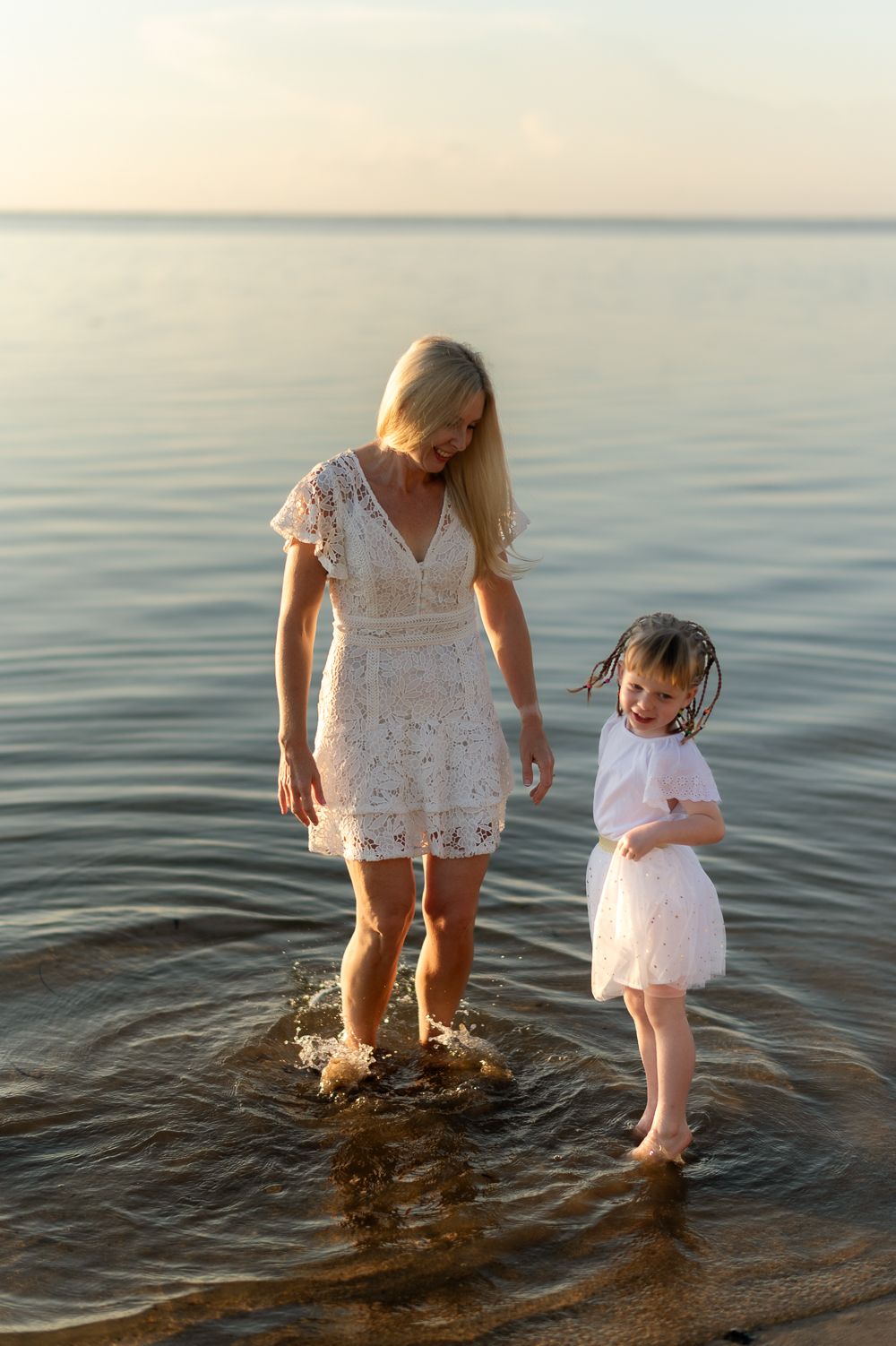 Mother and young daughter splashing in shallow ocean water during a warm golden sunset.