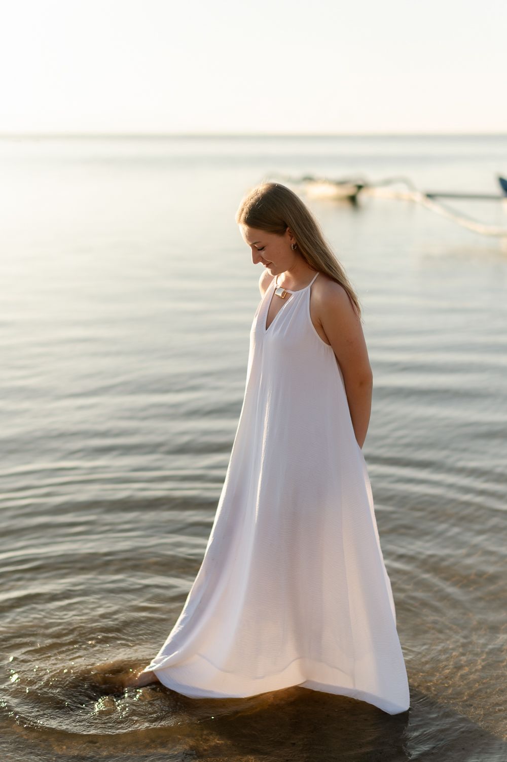 Woman in flowing white maxi dress wades through calm shallow ocean water at sunset.