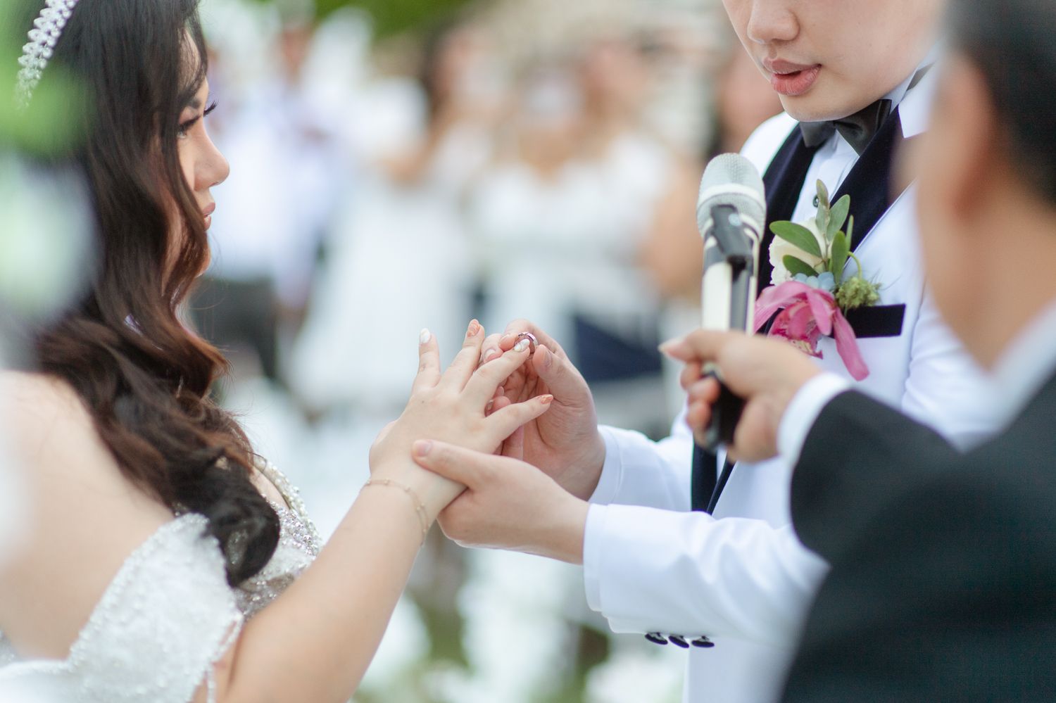 Bride and groom exchanging rings during an outdoor wedding ceremony, with officiant holding microphone.