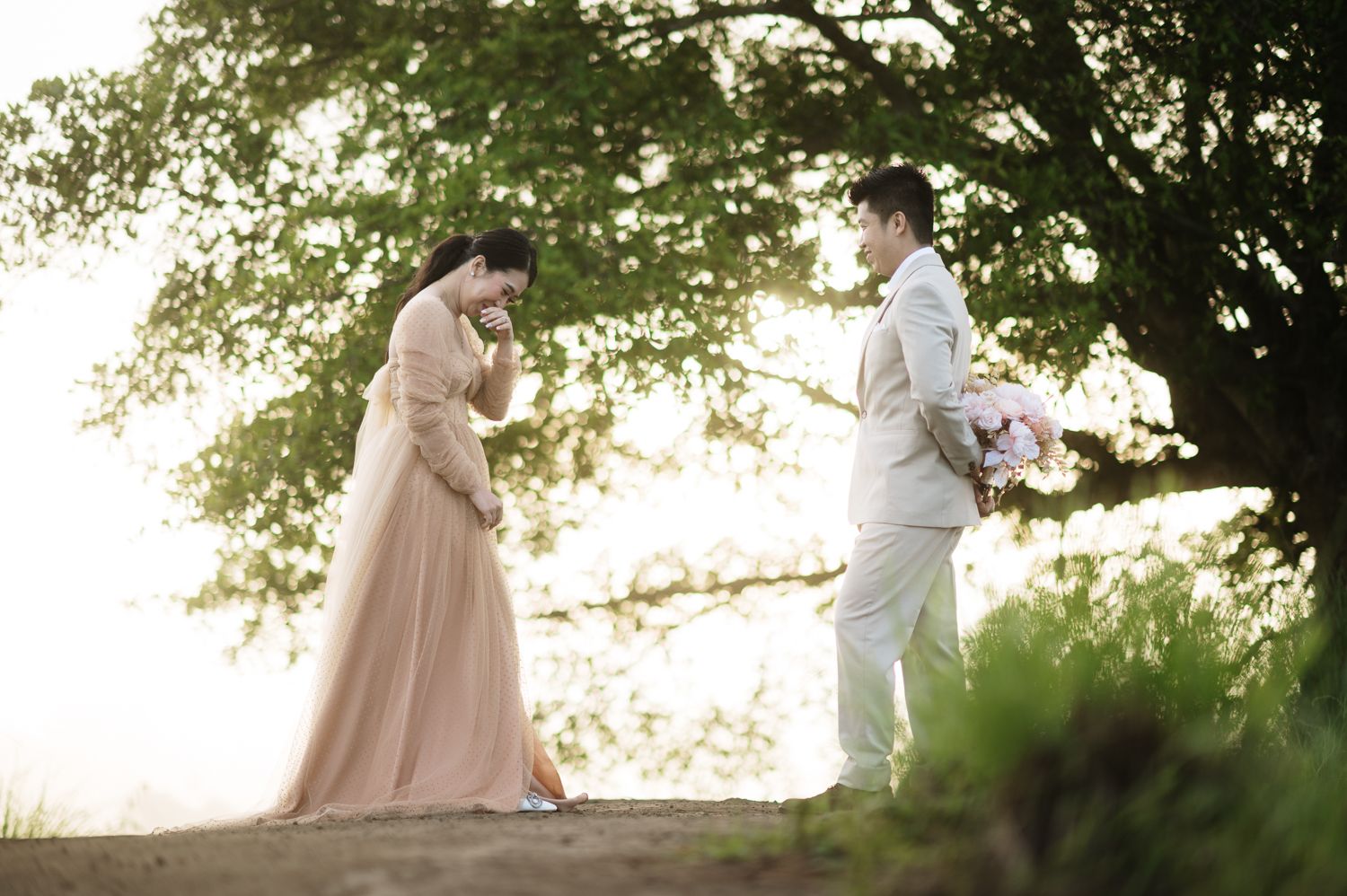 Couple in elegant wedding attire standing under a tree in golden light, bride in blush gown, groom holding bouquet.