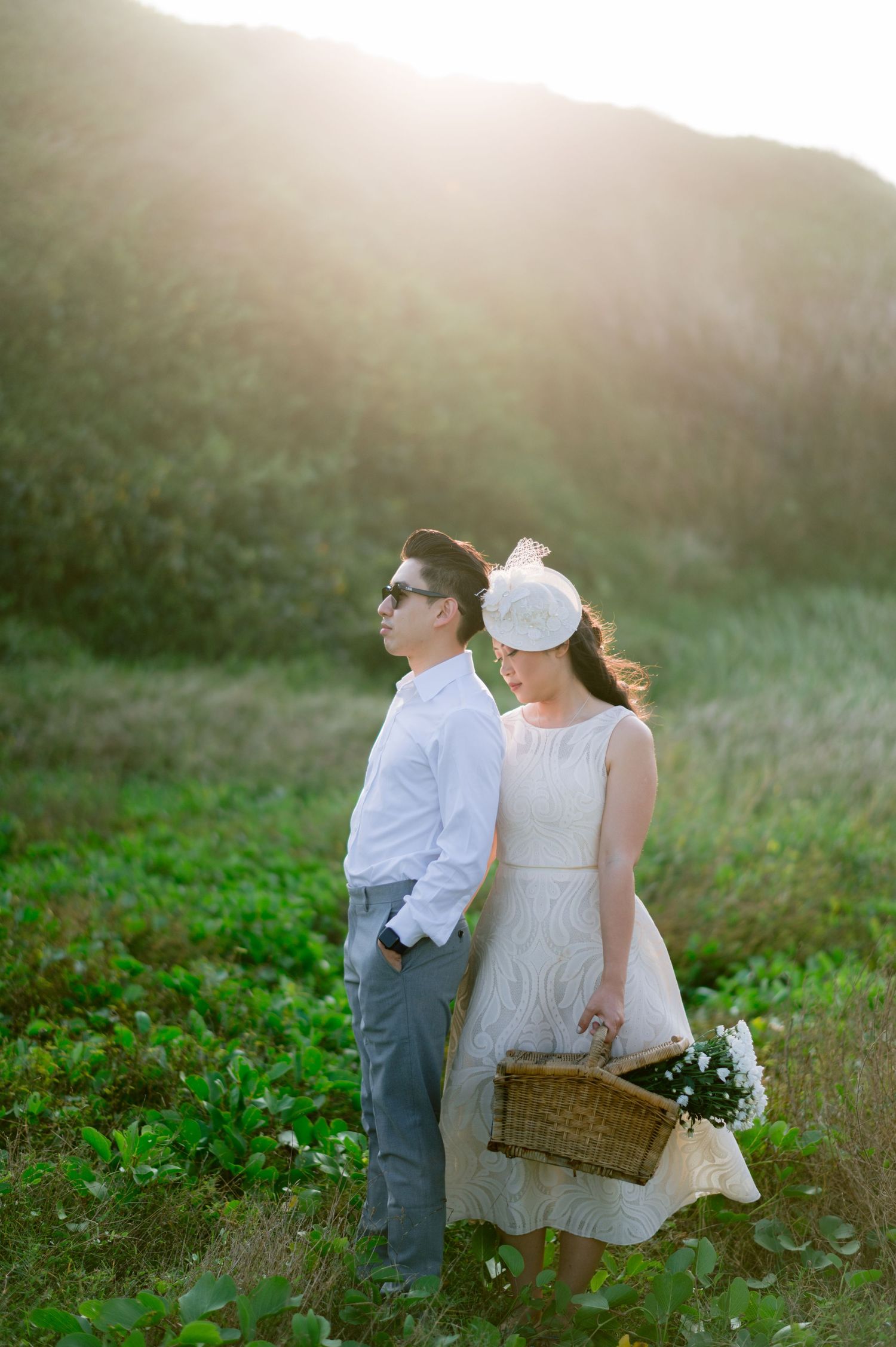 Couple in white attire stands in a sunlit green field, woman holding a wicker basket, golden light glowing behind hills.