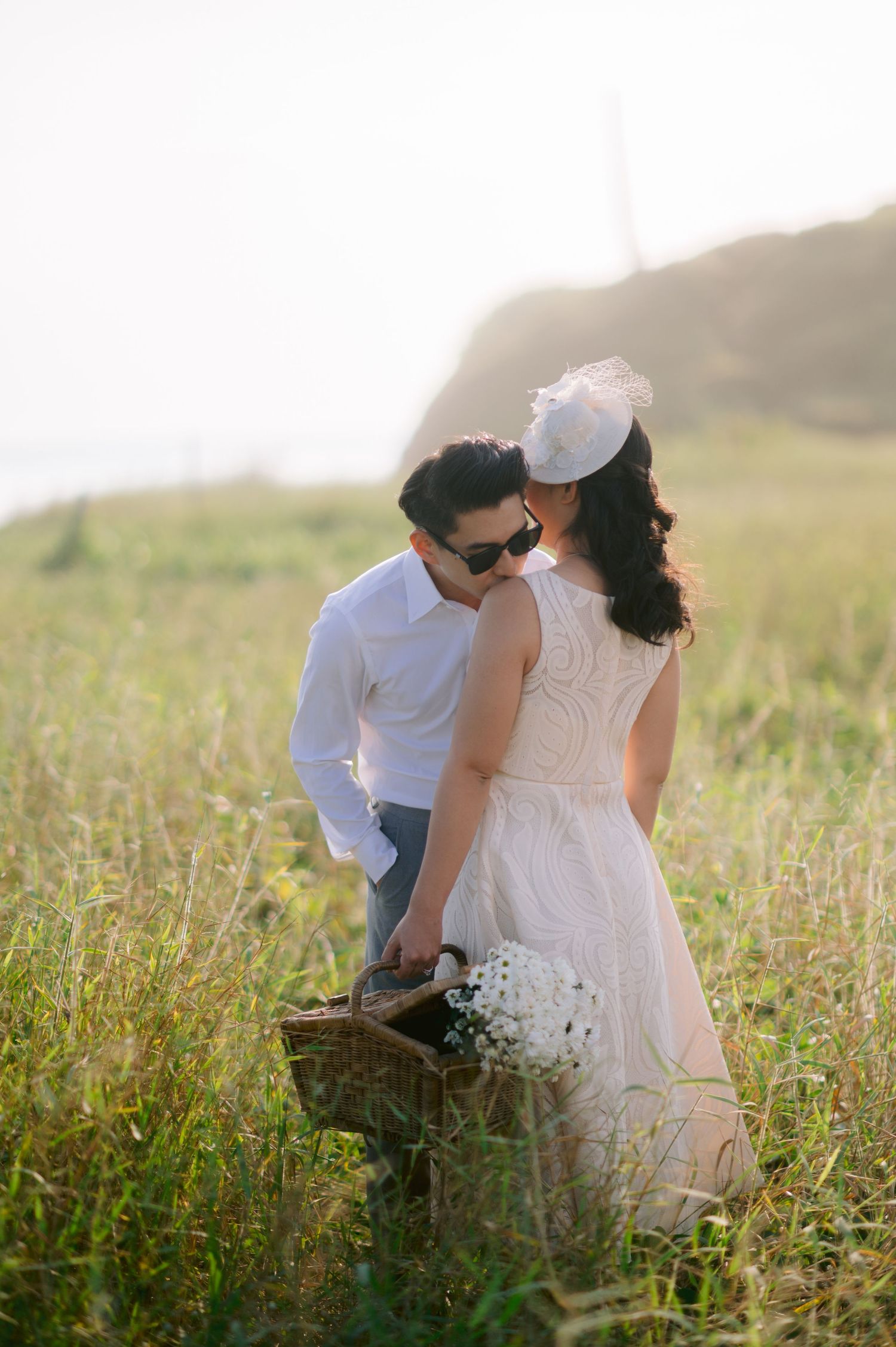 Couple sharing a romantic moment in a sunlit meadow, woman in white lace dress holding flowers, man in sunglasses with picnic basket.