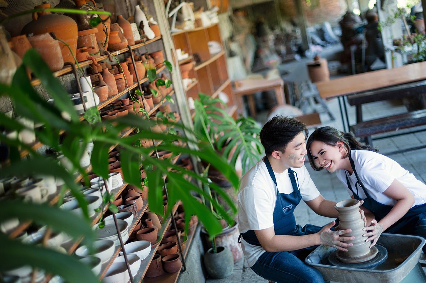 Couple smiling while shaping clay on a pottery wheel in a rustic ceramic shop surrounded by terracotta pots.