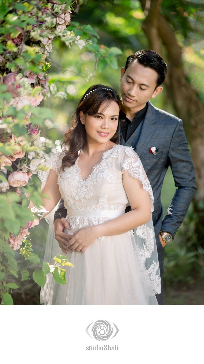 A couple poses romantically under floral arch, bride in lace gown and groom in gray suit, surrounded by pink blooms.