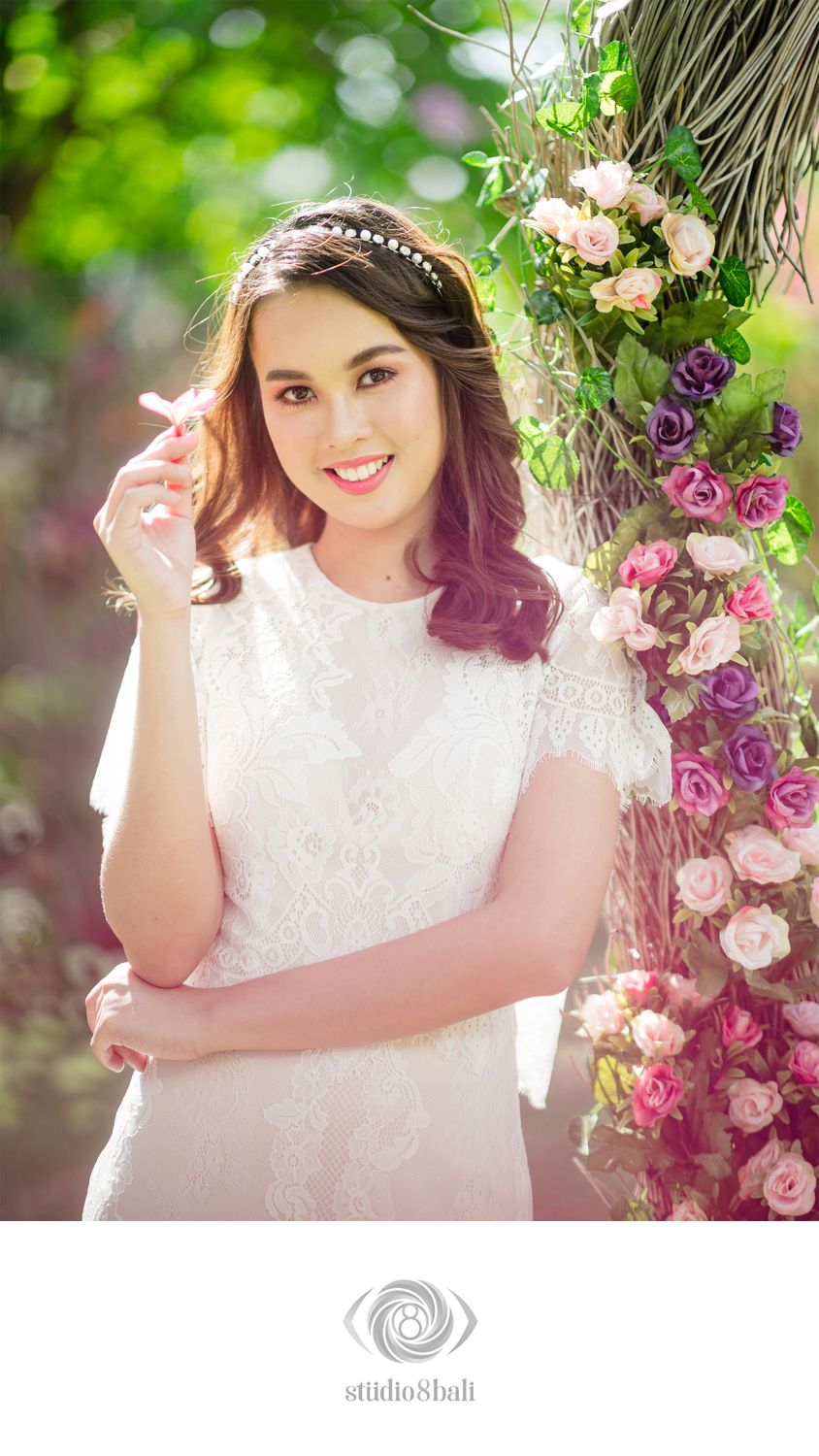 Smiling bride in white lace dress with floral headband poses beside pink and purple roses in a sunlit garden.