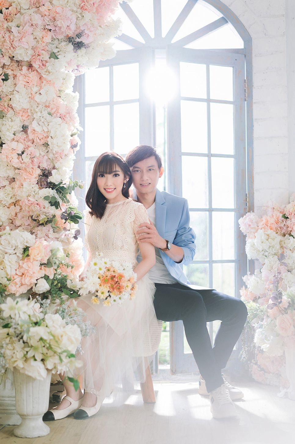 Couple posing in a floral studio with pink and white blooms, woman in white dress holding bouquet, man in blue blazer.