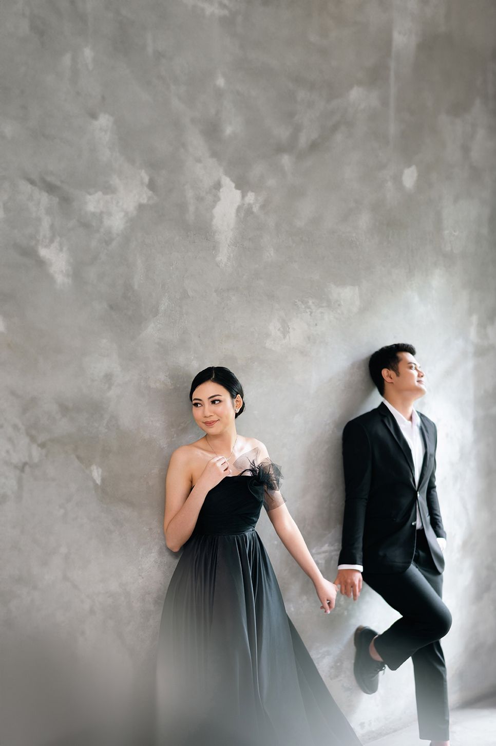 Elegant couple in matching black formal attire posing against a gray concrete wall in a stylish pre-wedding photo shoot.