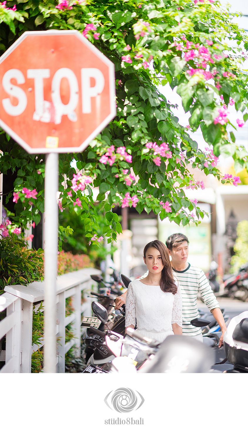 Couple posing near a stop sign surrounded by pink bougainvillea flowers and parked motorcycles on a sunny street.