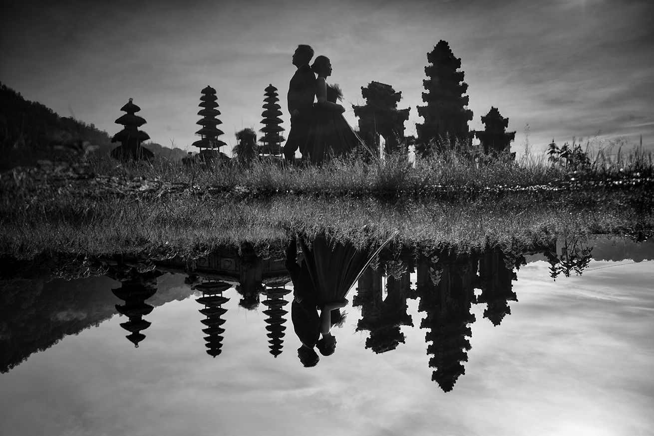 Black and white reflection of Balinese temple spires and silhouetted figures mirrored in still water.
