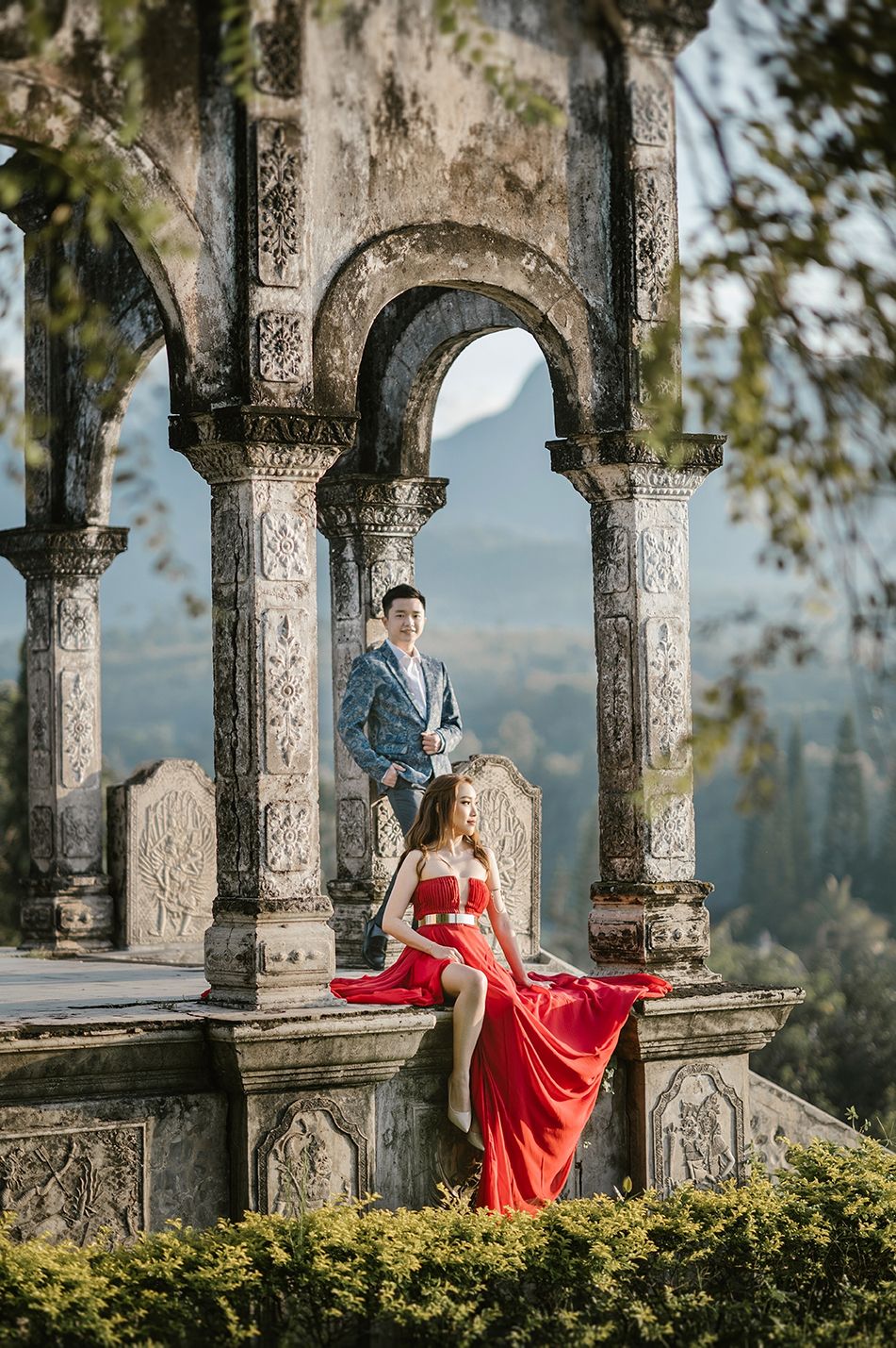Couple in red gown and blue suit posing at ornate stone archway with mountain views in Bali.