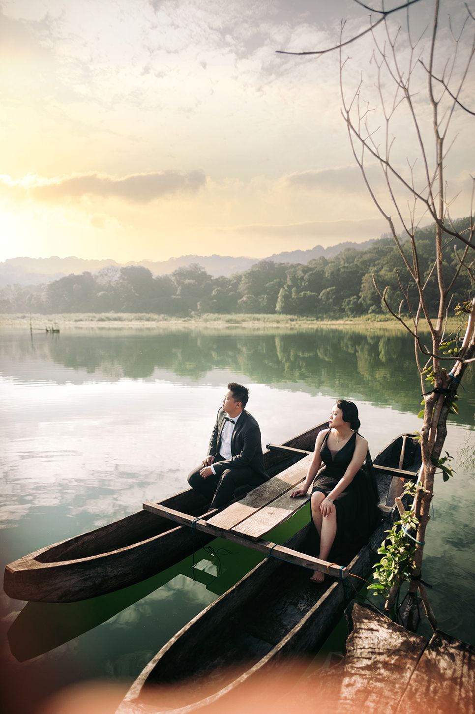 Couple in elegant attire sits in rustic wooden boat on calm lake at golden sunset with lush green hills.