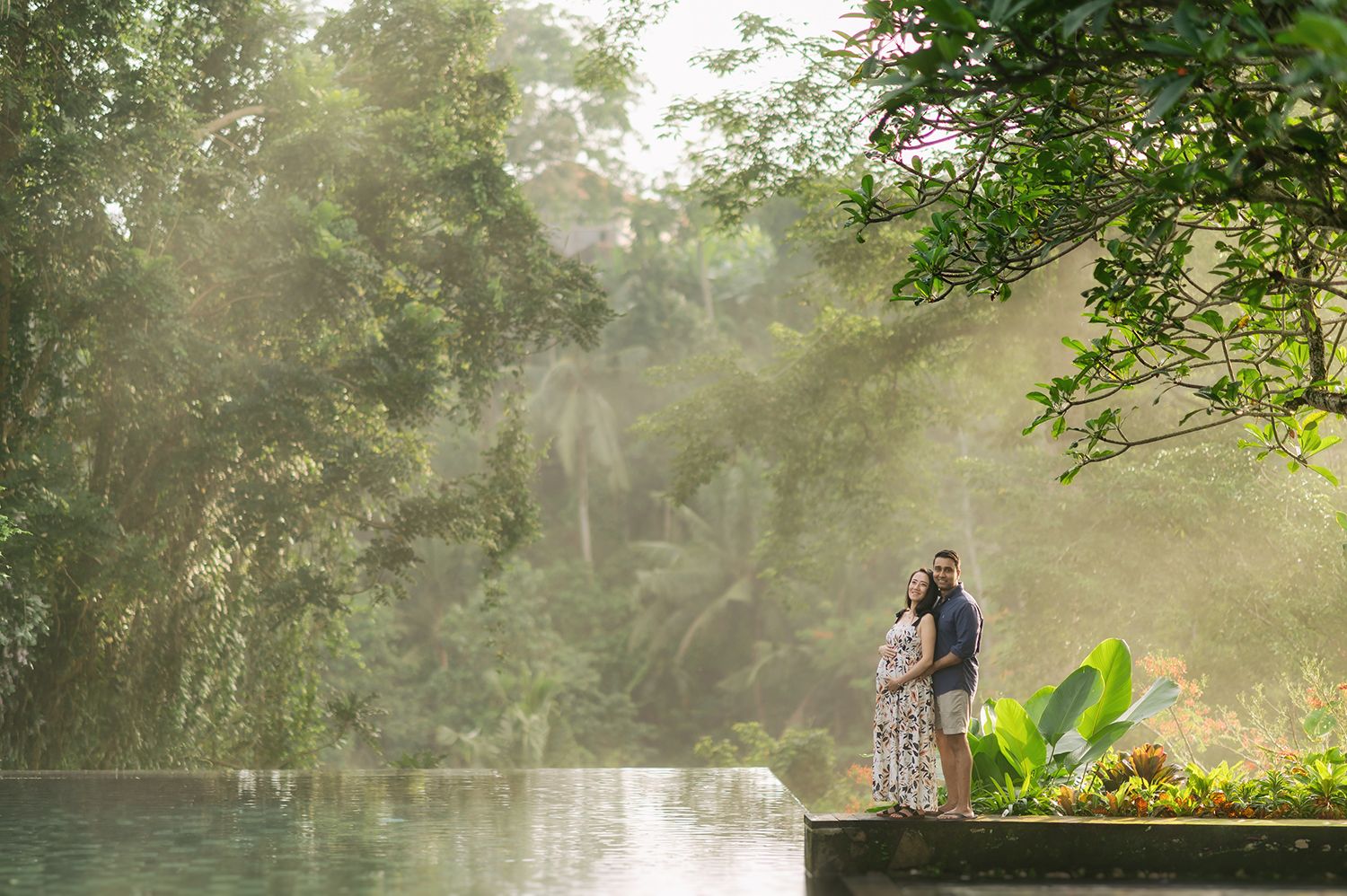 Couple embracing by a misty jungle infinity pool surrounded by lush tropical greenery and soft sunlight rays Maya Ubud Resort and Spa in Ba.