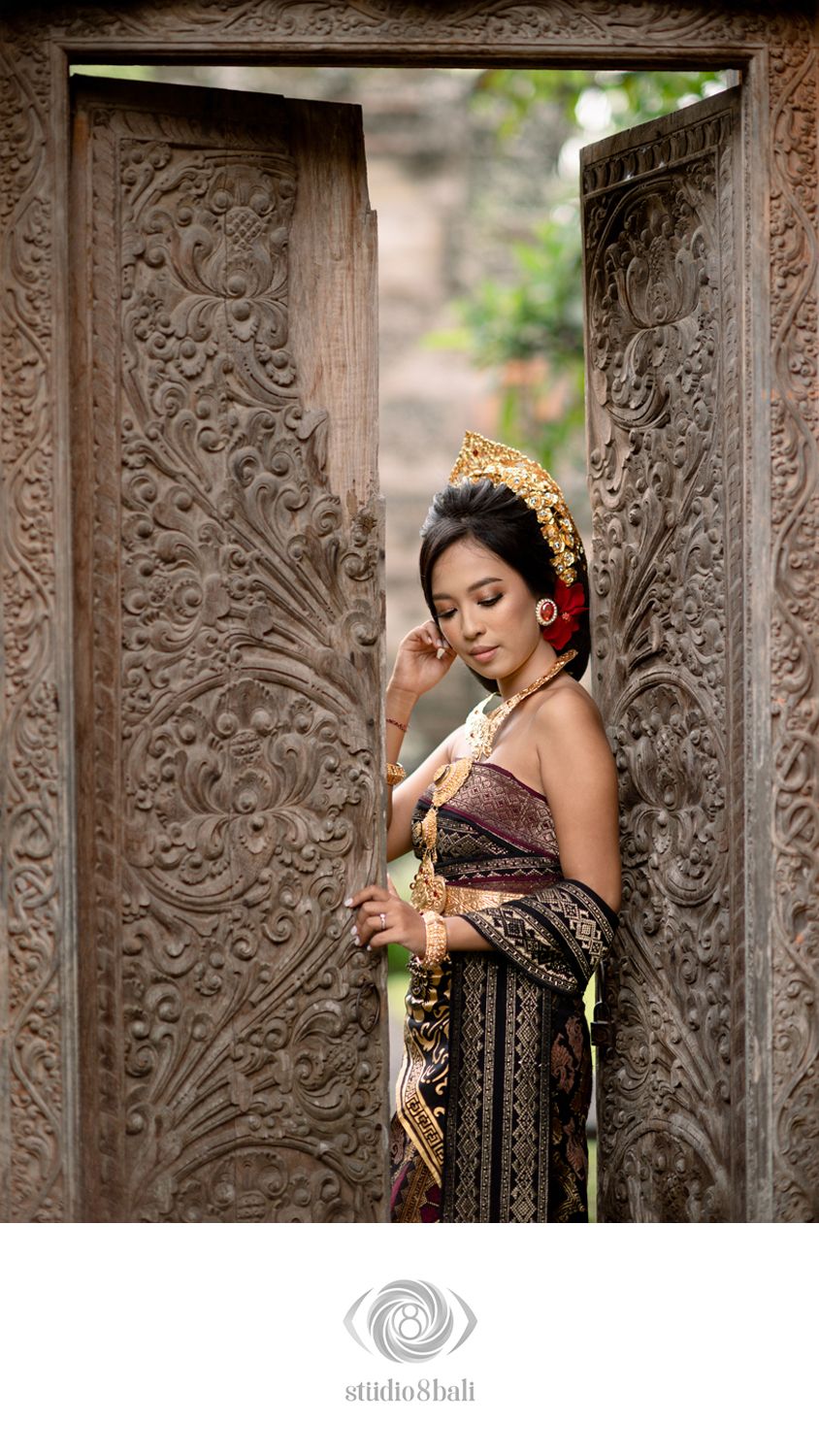 Balinese woman in traditional gold kebaya and headdress poses gracefully between ornate carved wooden temple doors.