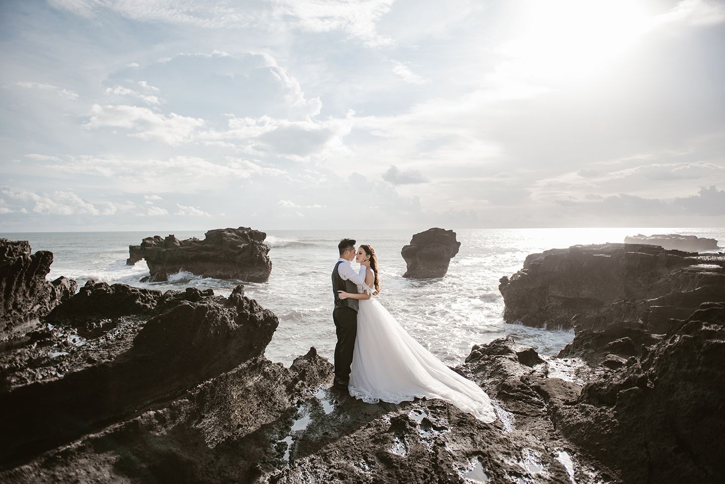 Bride and groom kissing on rocky ocean cliffs with dramatic waves and sunlit sky in a stunning wedding photo.